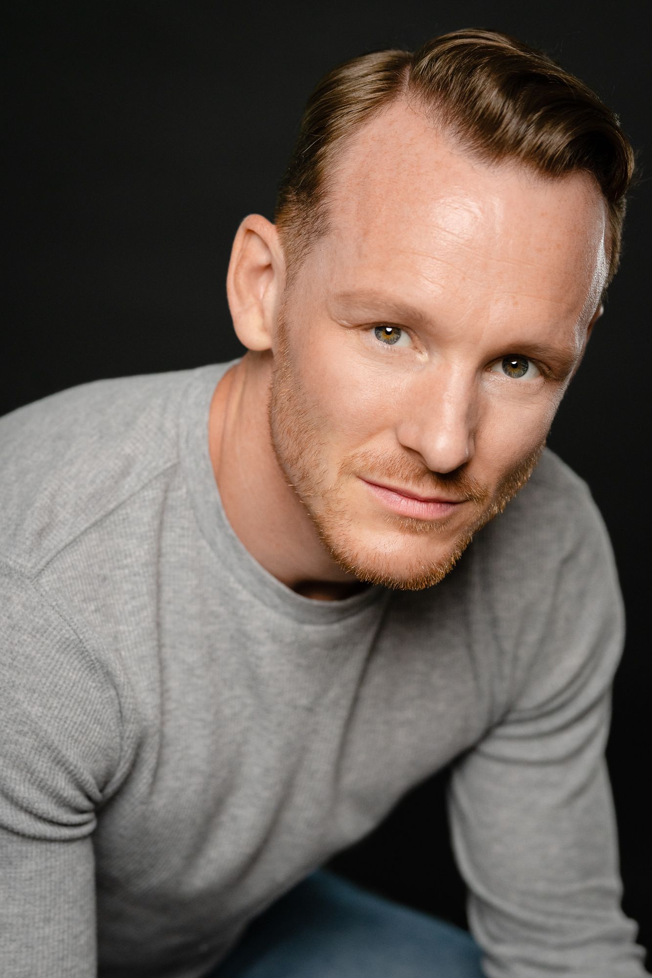 A red-haired man in a gray sweater poses confidently against a dark background in a professional headshot.