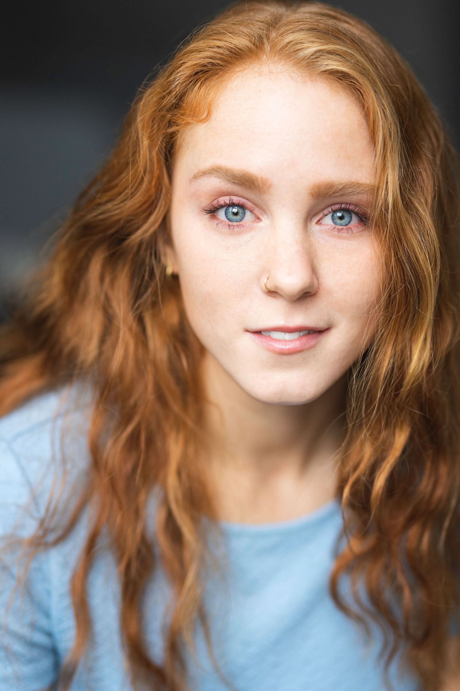 Young woman with long red wavy hair, blue eyes, freckles, and a light blue top smiles softly at the camera.