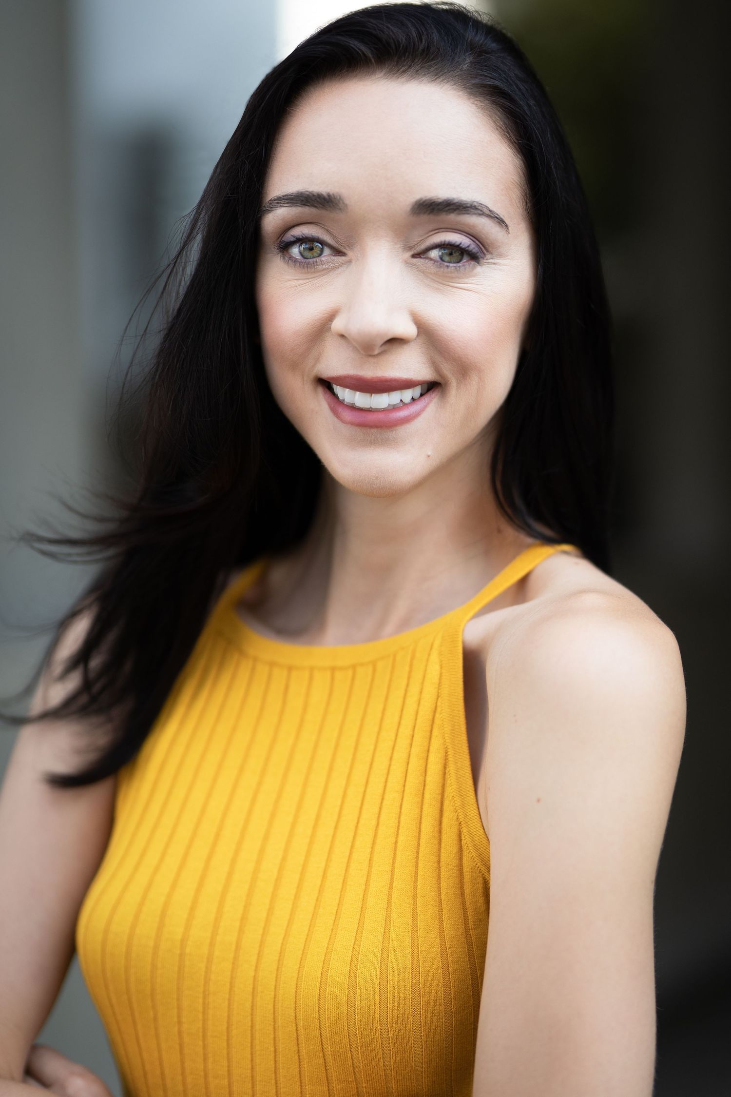 Smiling woman with long dark hair wearing a yellow ribbed sleeveless top, posing confidently outdoors.