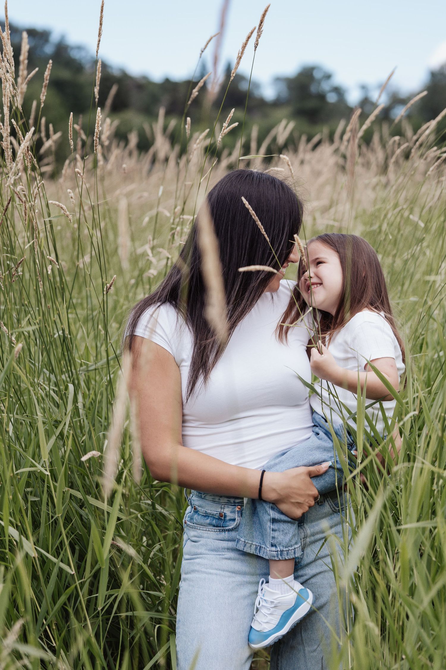 Mother and daughter in white shirts and jeans embrace in a tall grass field, sharing a joyful moment outdoors.