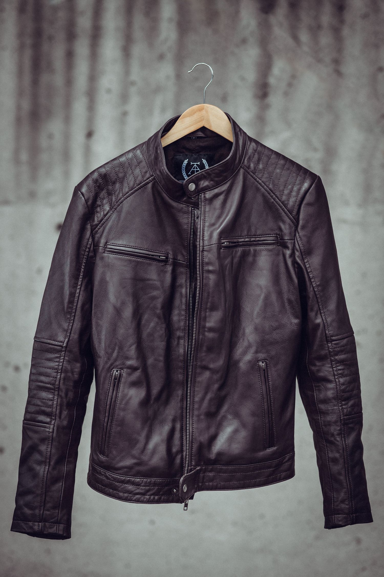 Dark brown leather biker jacket on wooden hanger against concrete wall, featuring zip front and quilted shoulder details. product photo