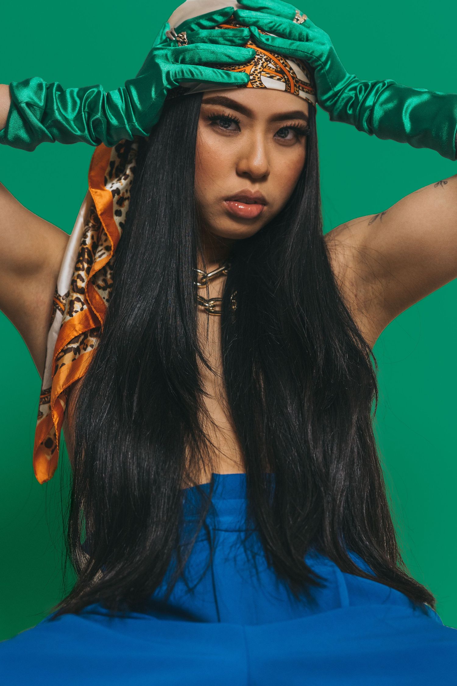 Young woman with long black hair wearing a blue top, green satin gloves, and a patterned headscarf on green background. Vancouver photo