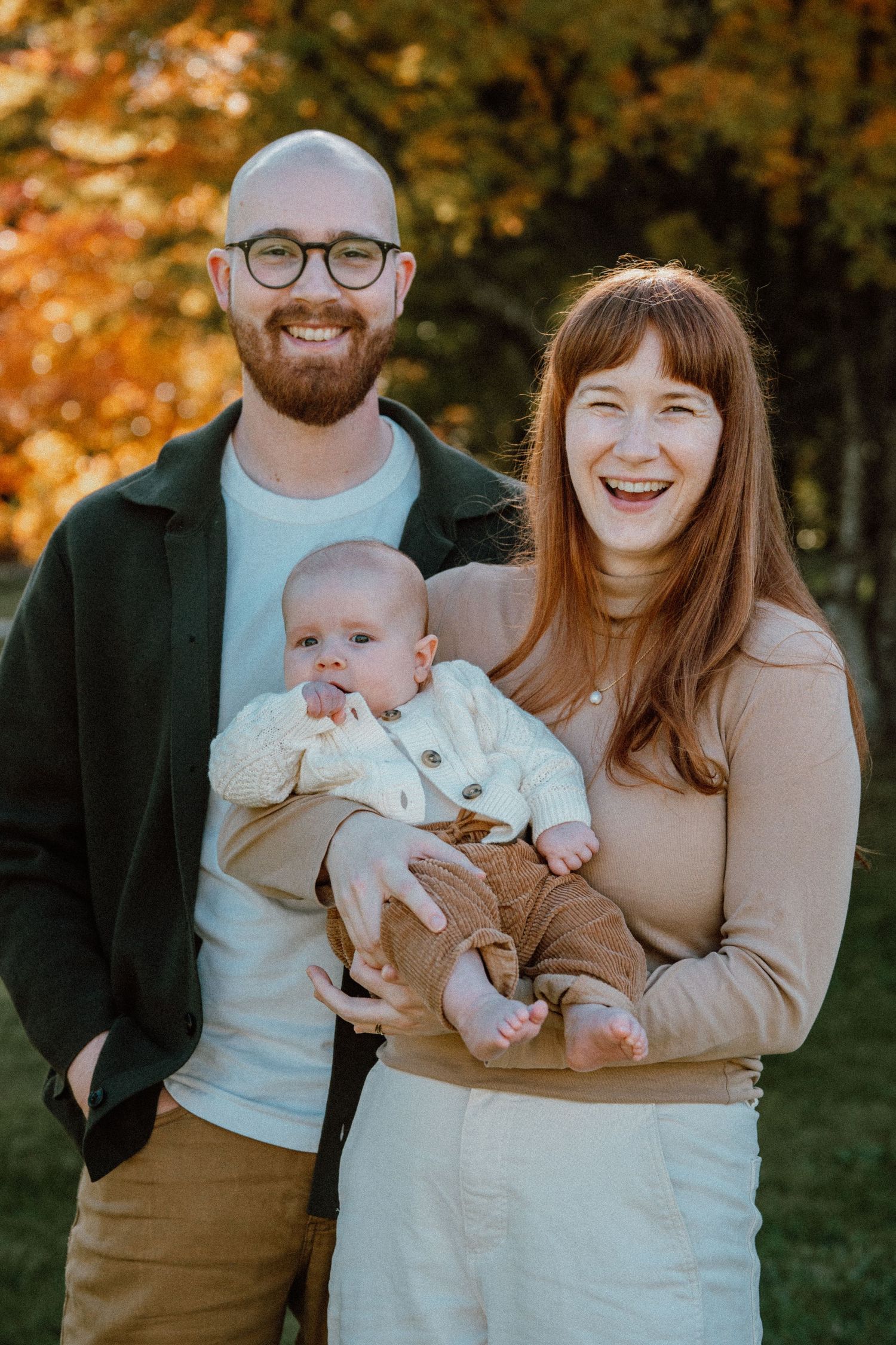 Happy couple smiling outdoors in autumn, holding their baby dressed in cream and brown, with colorful fall foliage behind.