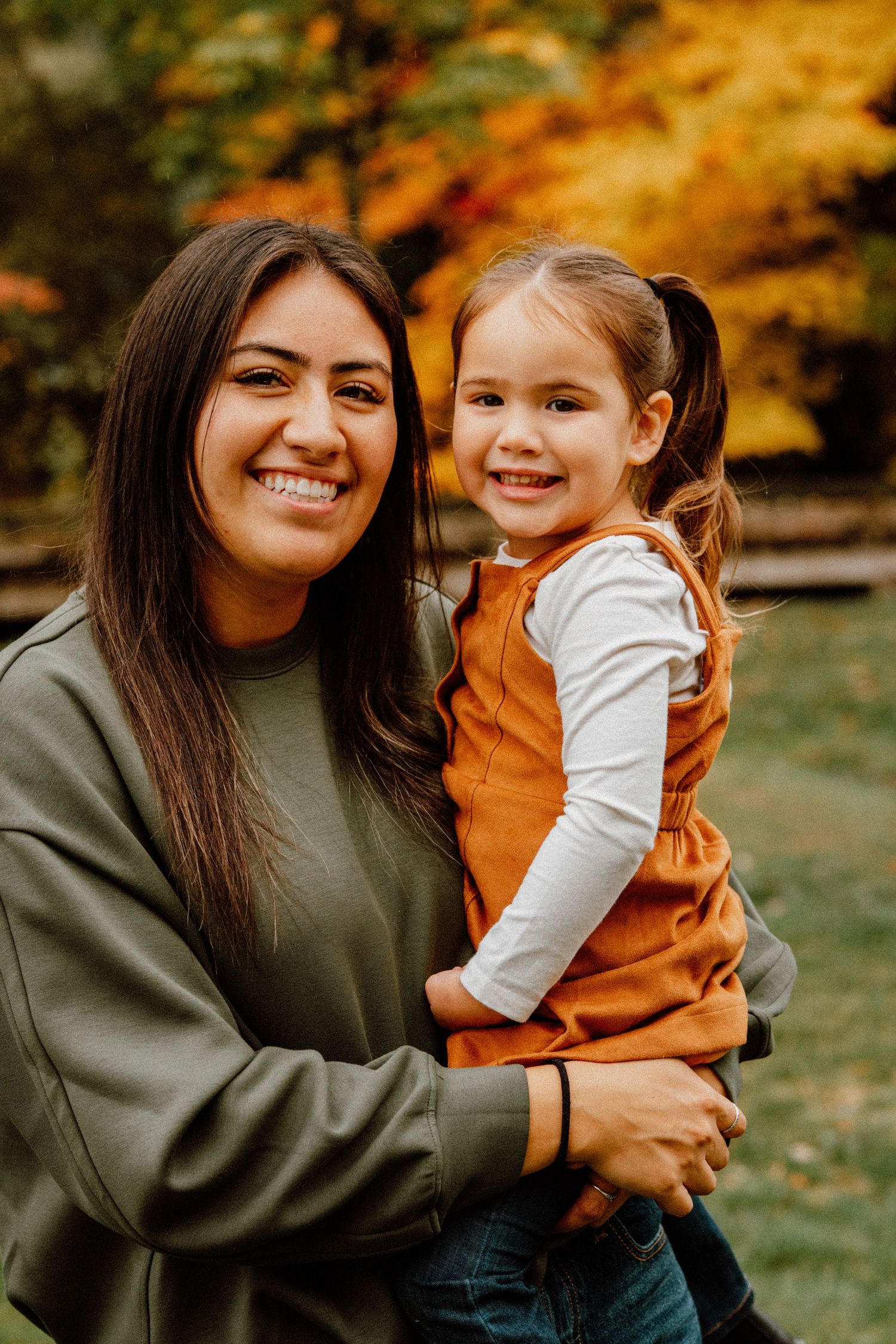 A smiling woman in a green sweatshirt holds a happy young girl in an orange dress outdoors during fall.