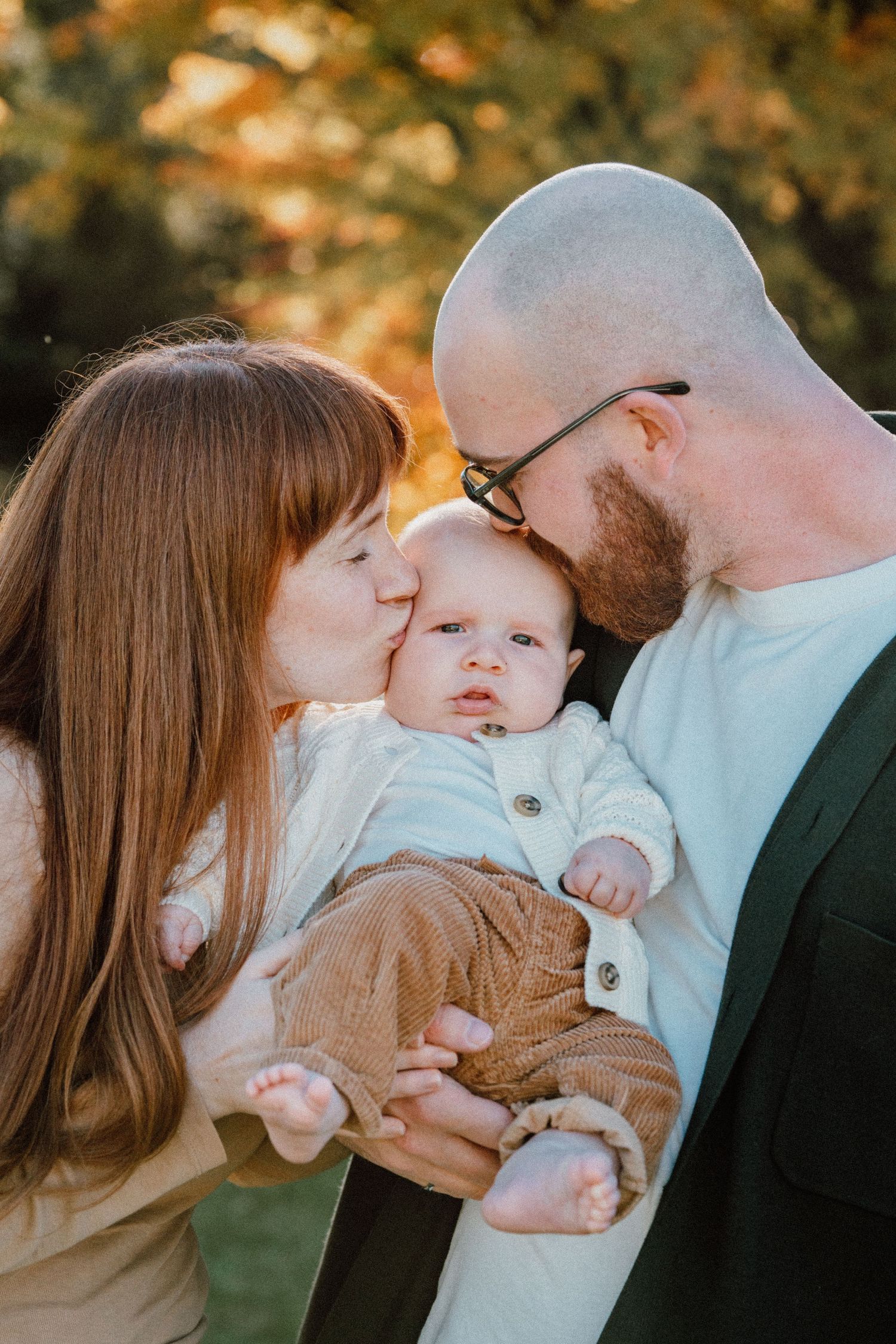 Two parents kissing their newborn baby on the cheeks outdoors in autumn light.