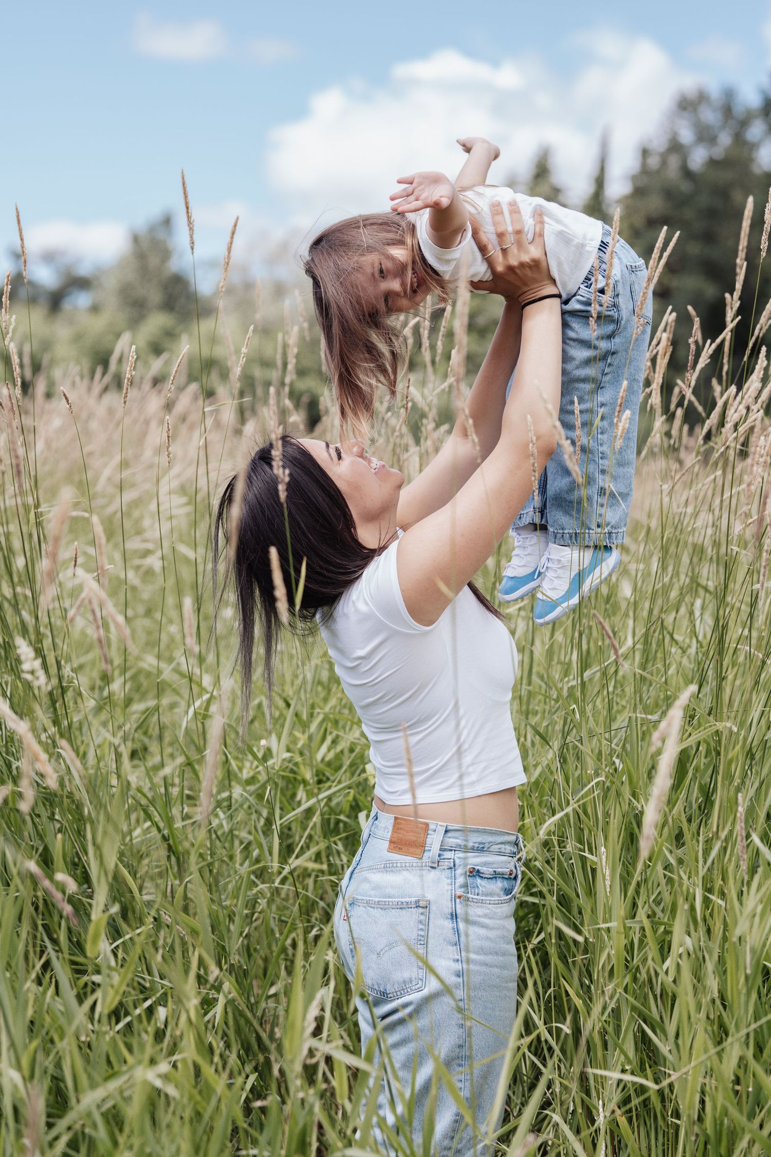 A mother joyfully lifts her toddler in a lush green field under a partly cloudy sky, both wearing jeans and white tops.
