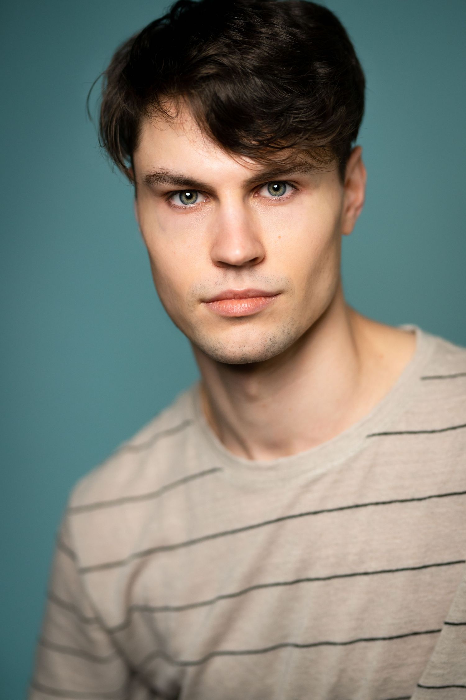 Young man with dark hair and blue eyes wearing a striped beige t-shirt posed against a teal background.