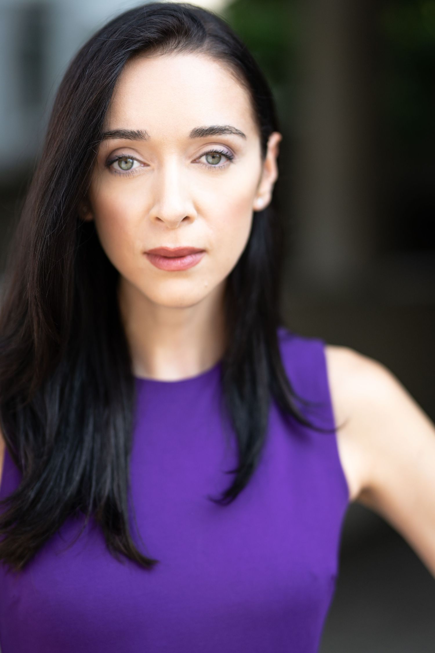Woman with dark hair in purple sleeveless top poses for professional headshot outdoors with blurred background.