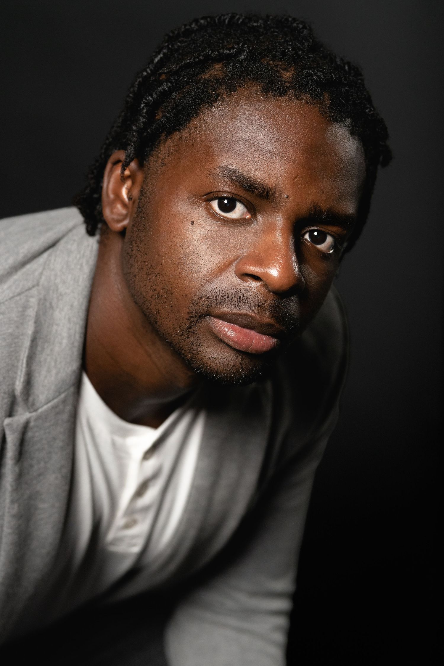 Close-up portrait of a young Black man with braided hair wearing a gray hoodie against a dark background.
