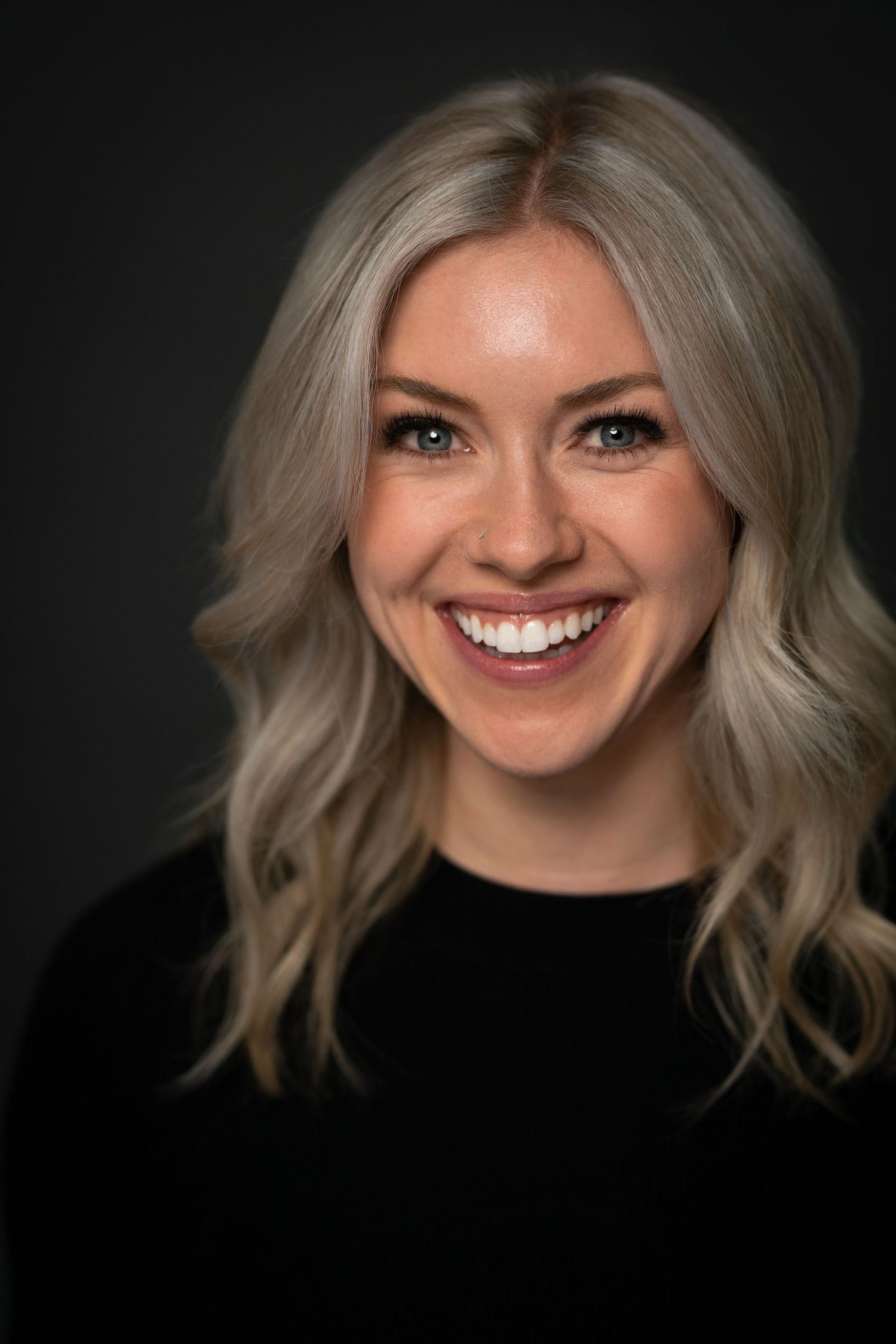 Smiling blonde woman with wavy hair wearing a black top against a dark background in a professional headshot.
