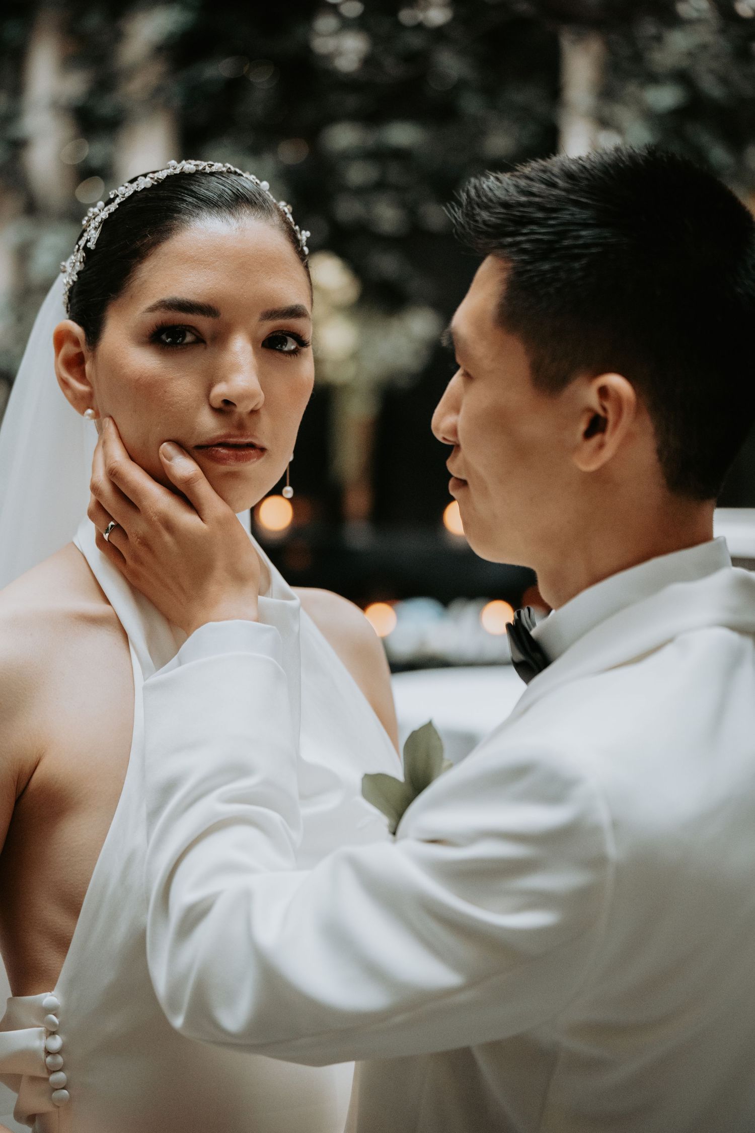 Groom tenderly cups bride's face during romantic outdoor wedding ceremony, both dressed in elegant white attire.