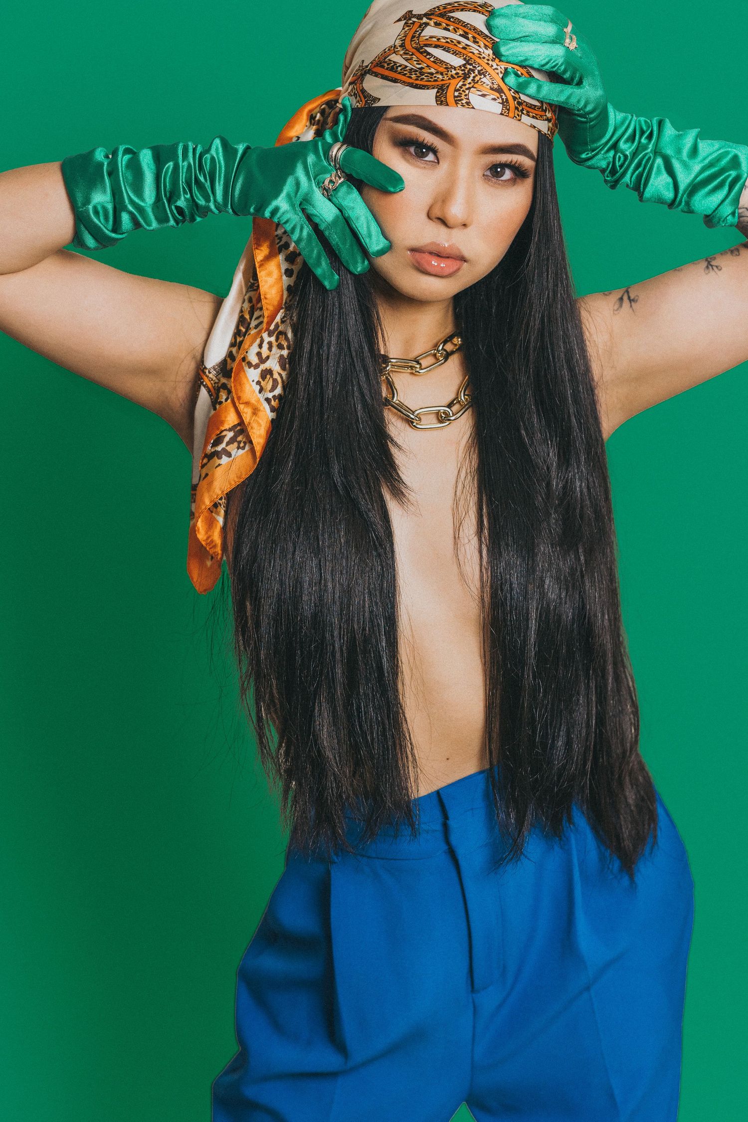 Woman with long black hair wearing green satin gloves, orange scarf headband, gold chain necklace, and blue pants on green background.