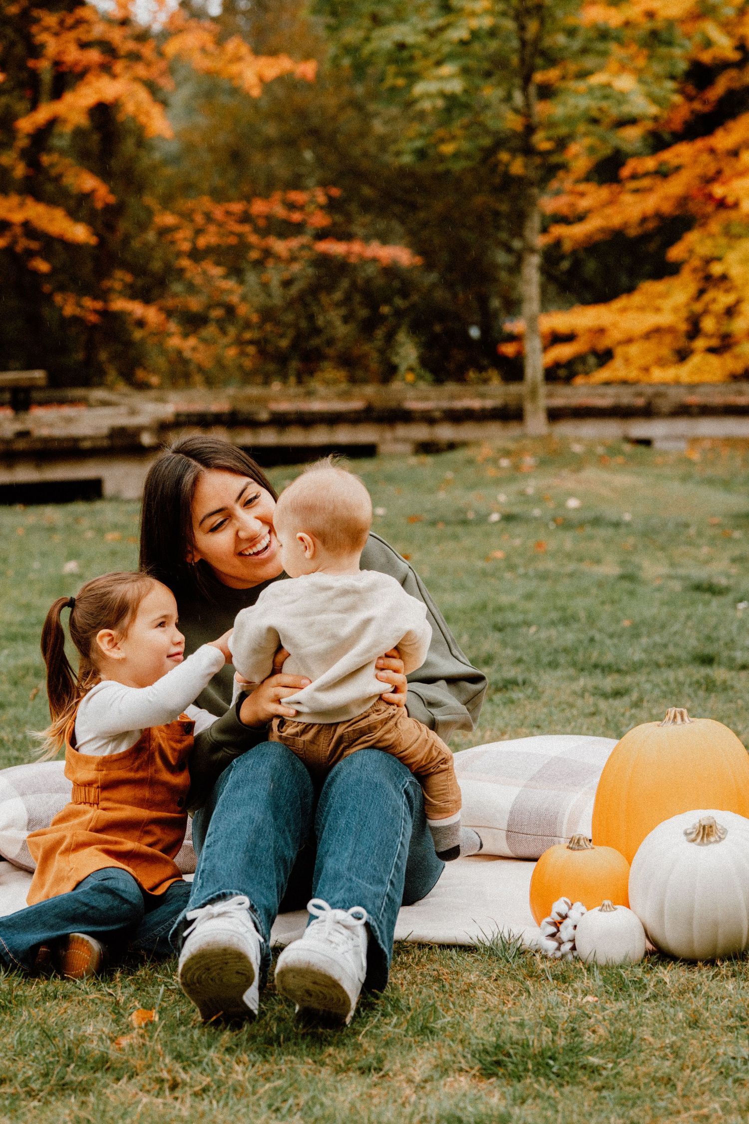 A mother sits on a blanket with her two young children surrounded by pumpkins and fall foliage in a park.