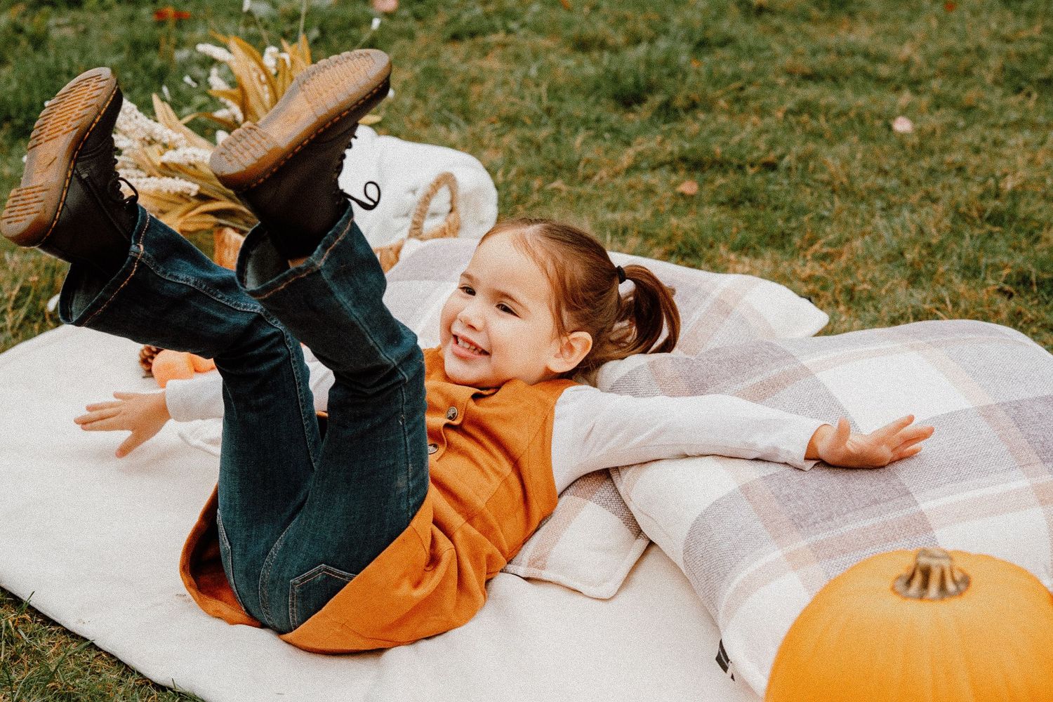 A happy young girl in an orange sweater plays on a plaid blanket surrounded by pumpkins in a fall outdoor setting.