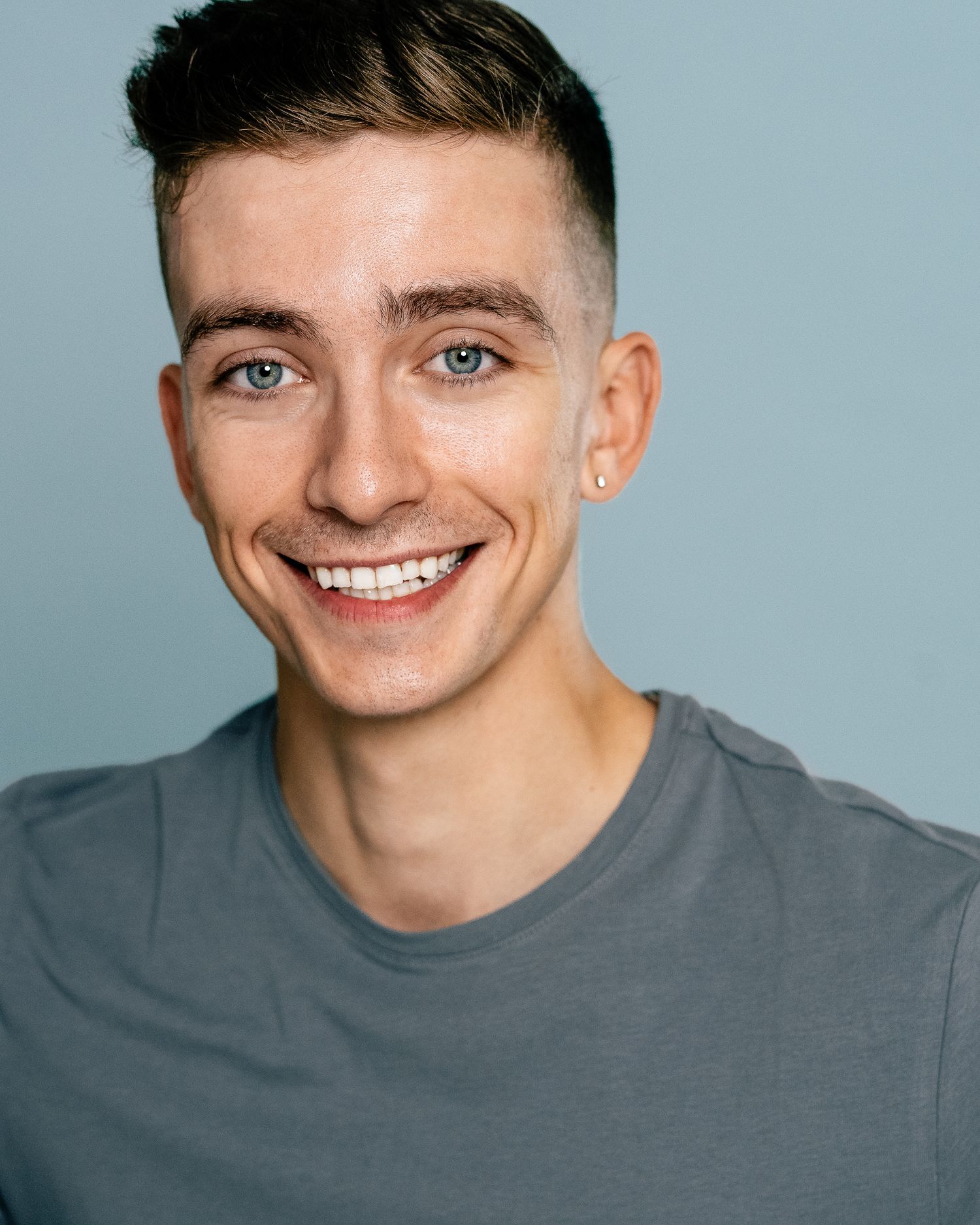 Young man with blue eyes and short brown hair smiling confidently in a gray t-shirt against a light blue background.