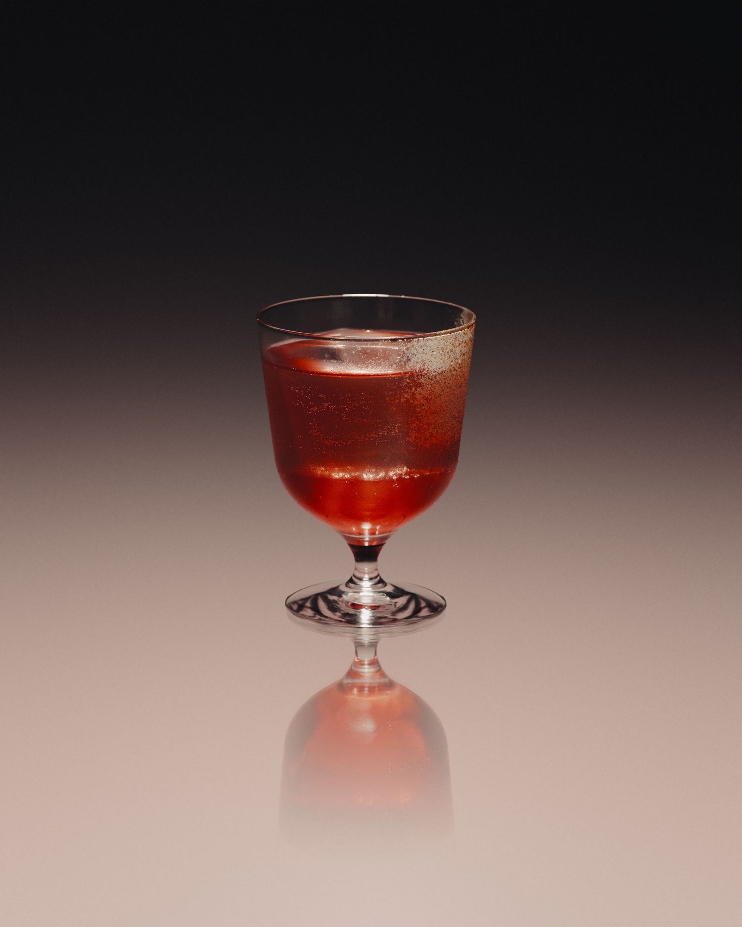 A deep red cocktail served in a classic footed glass, reflecting on a smooth surface against a dark gradient background.