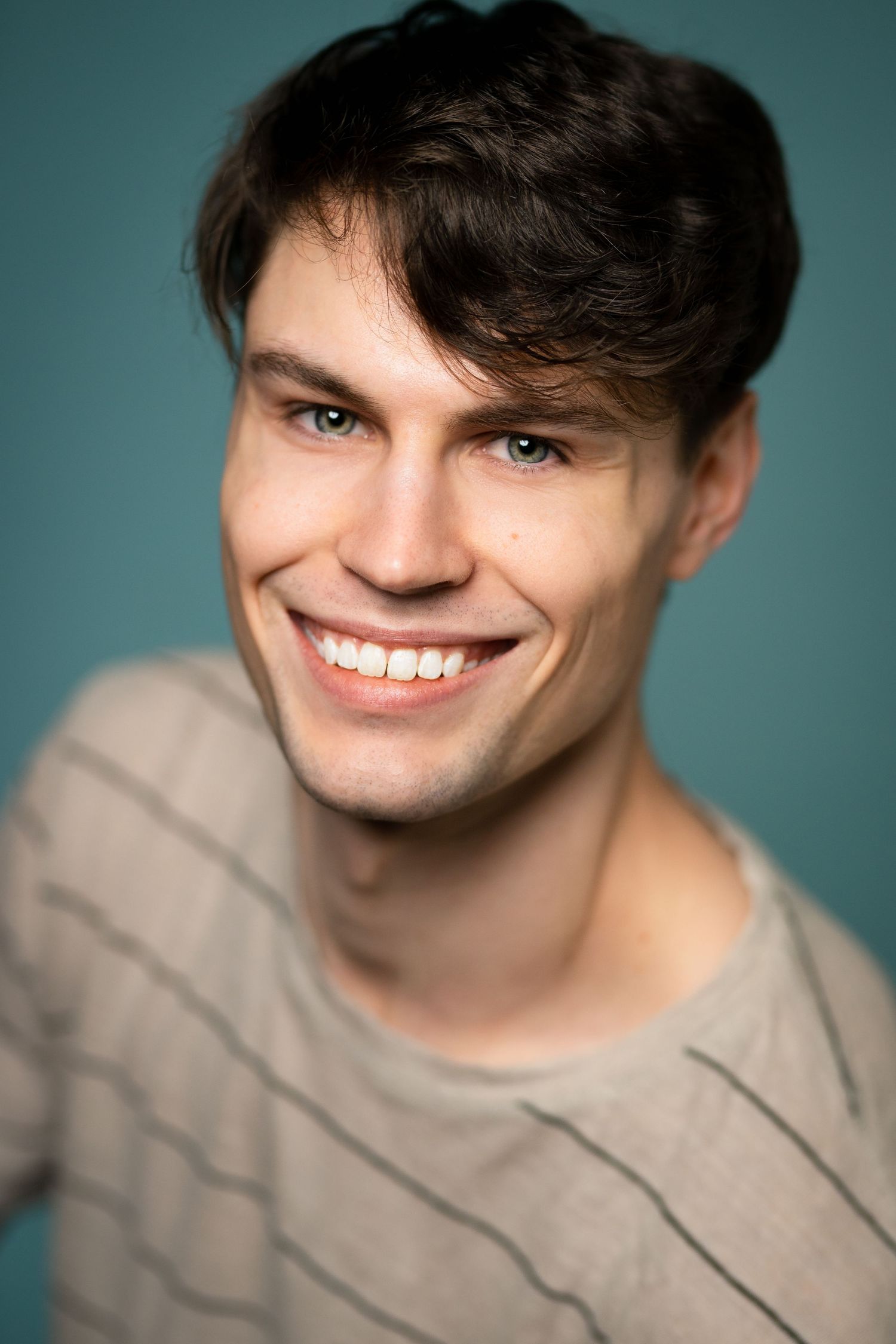 Young man with dark hair and blue eyes smiling broadly, wearing a striped sweater against a teal background.