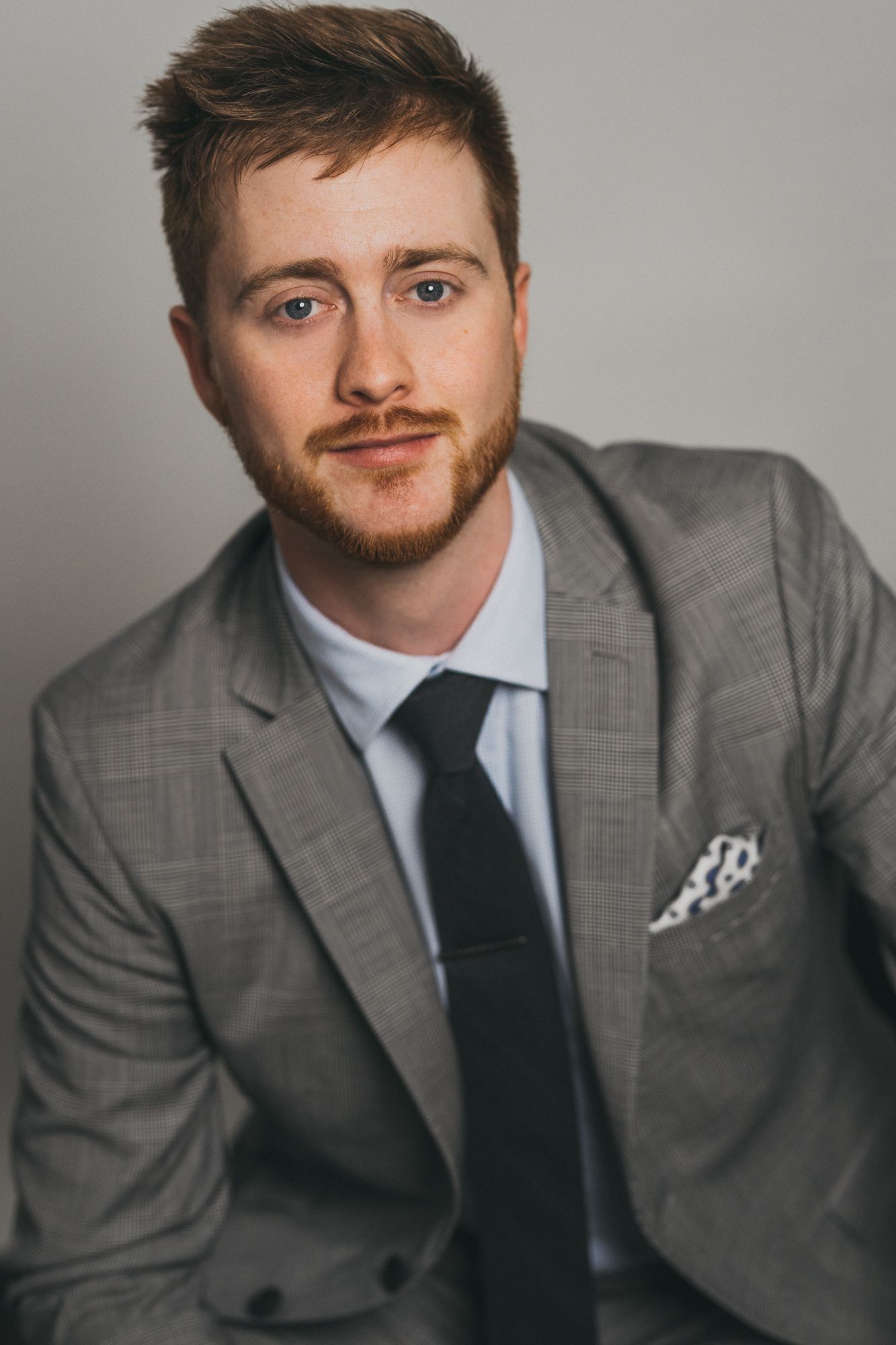 Young professional man with red beard wearing gray plaid suit, black tie, and white pocket square against gray backdrop.