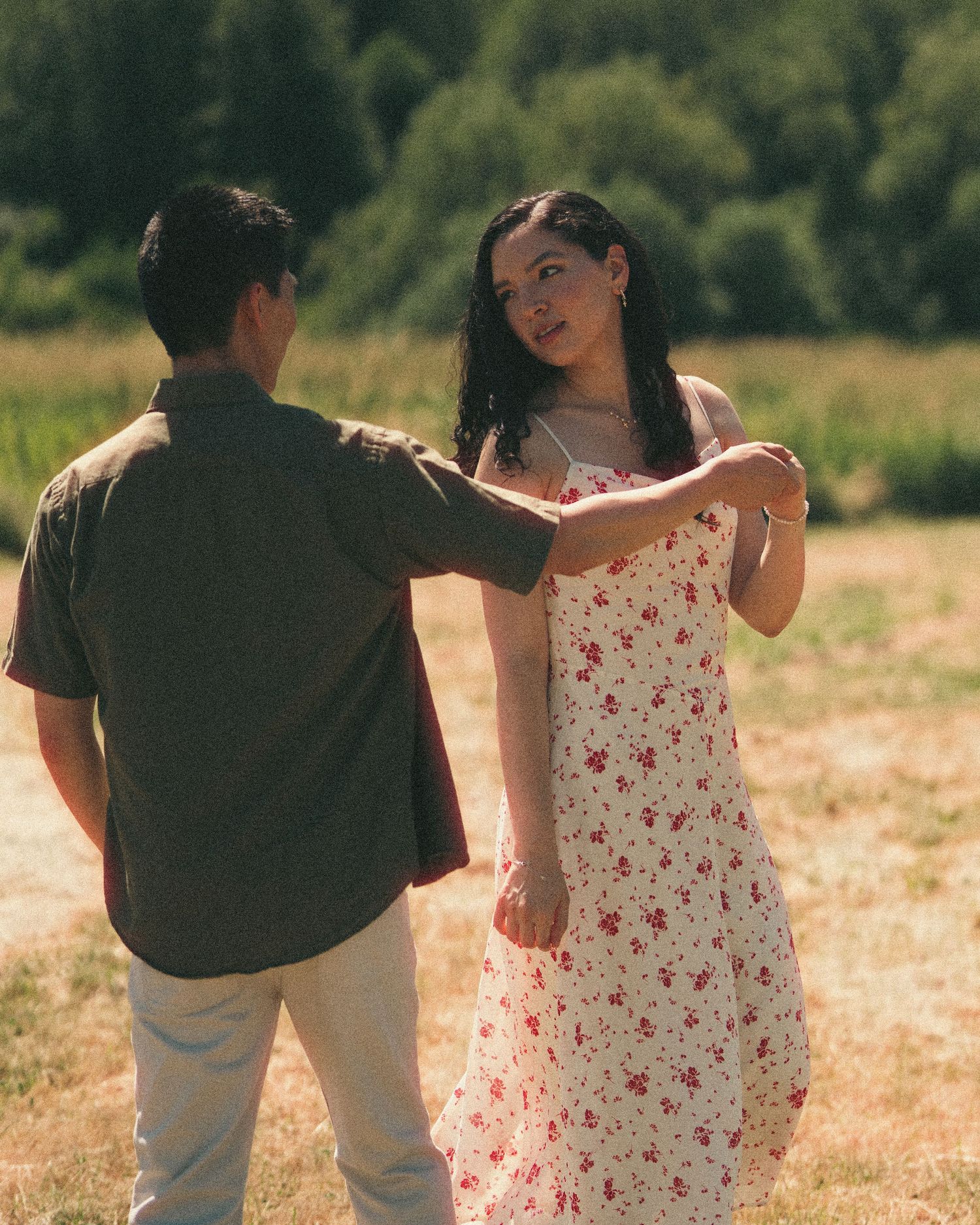 Couple dancing in a sunny field, woman in floral sundress holding hands with man in olive shirt.