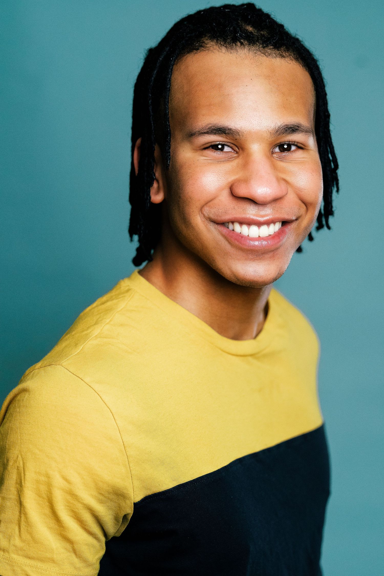 Smiling young man with dreadlocks wearing a yellow and black t-shirt against a teal background.