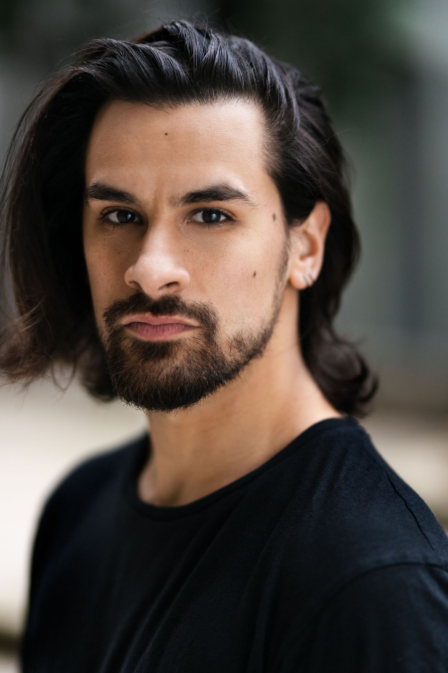 Young man with long dark hair, beard, and ear piercing wearing a black t-shirt, posing outdoors.