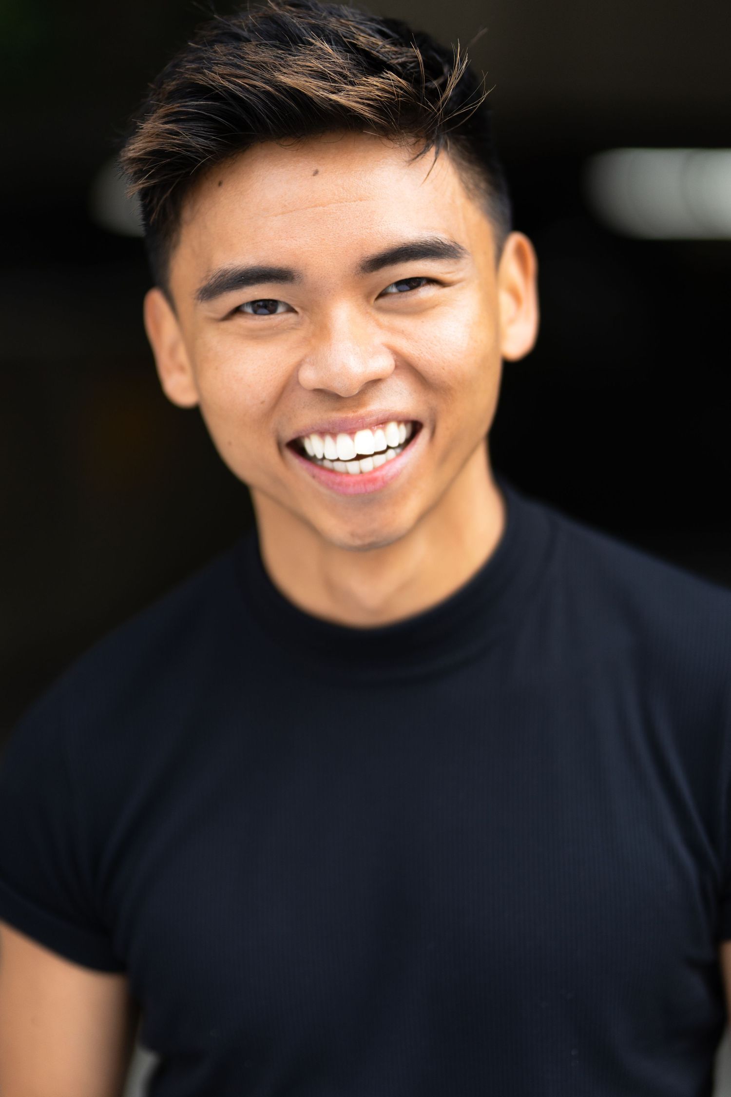 Young Asian man smiling broadly in a black t-shirt against a dark background.