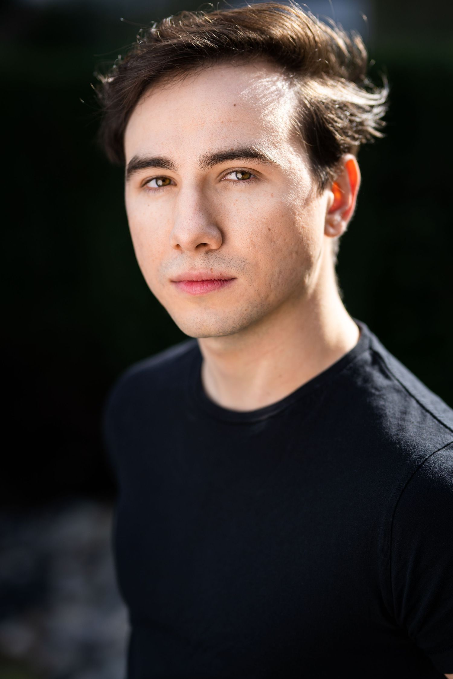 Young man with dark hair wearing a black t-shirt poses for an outdoor portrait with blurred green background.