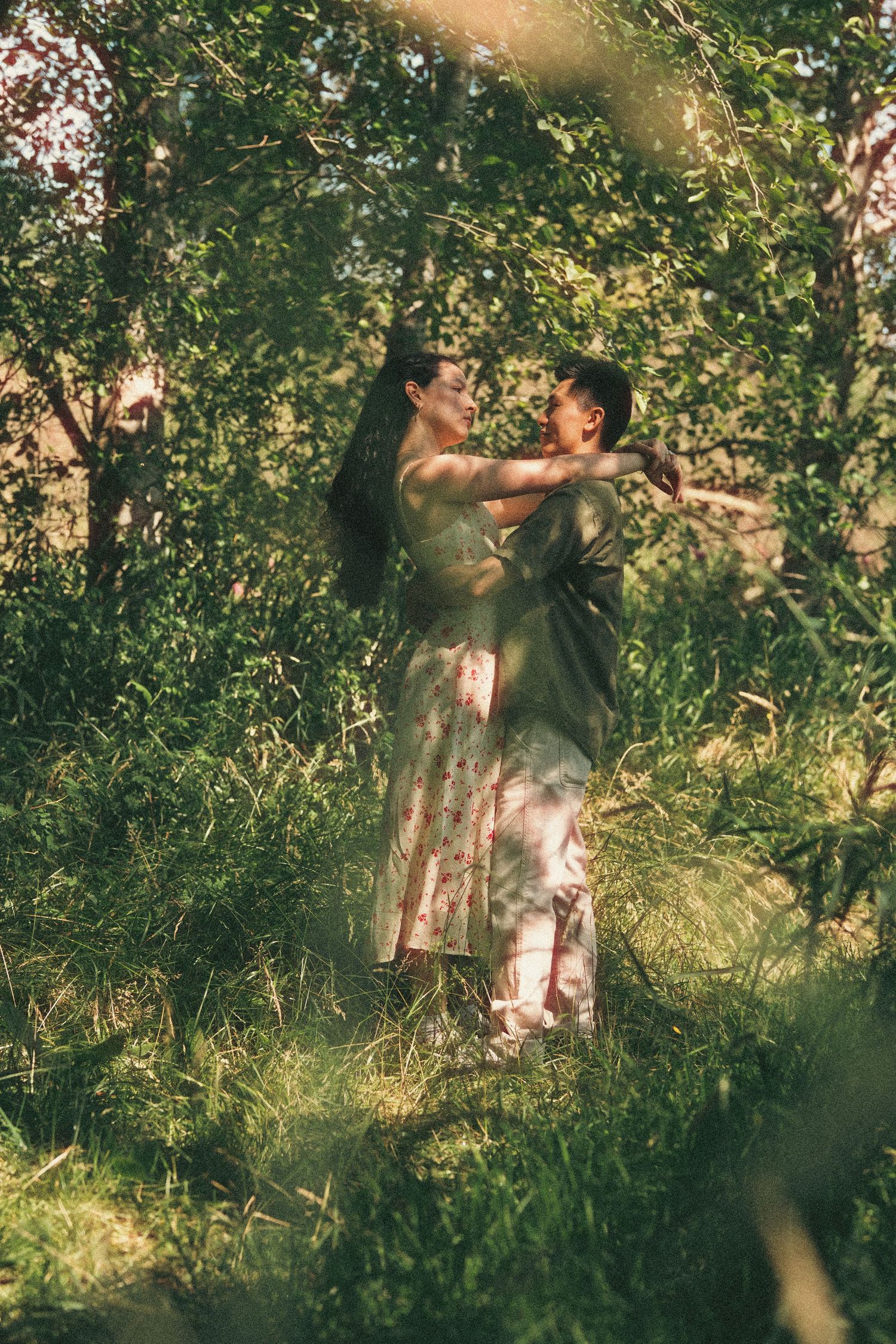 Couple embracing romantically in a lush green forest, surrounded by trees and tall grass in soft natural light.