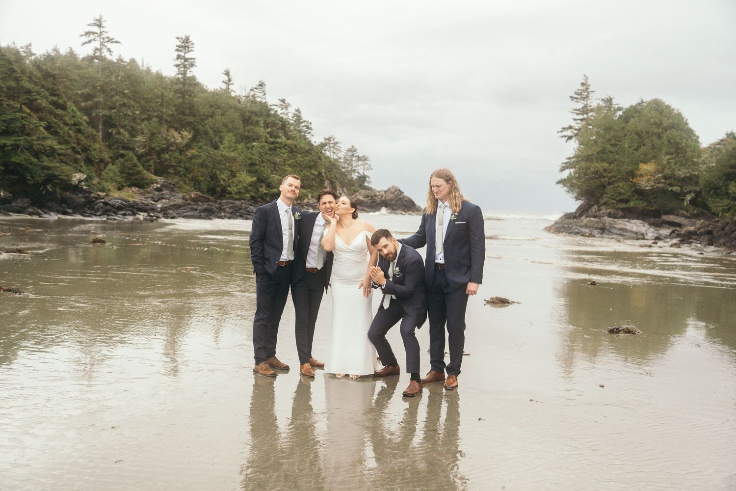 Wedding party posing on a scenic Pacific Northwest beach with rocky shores and evergreen trees in the background.