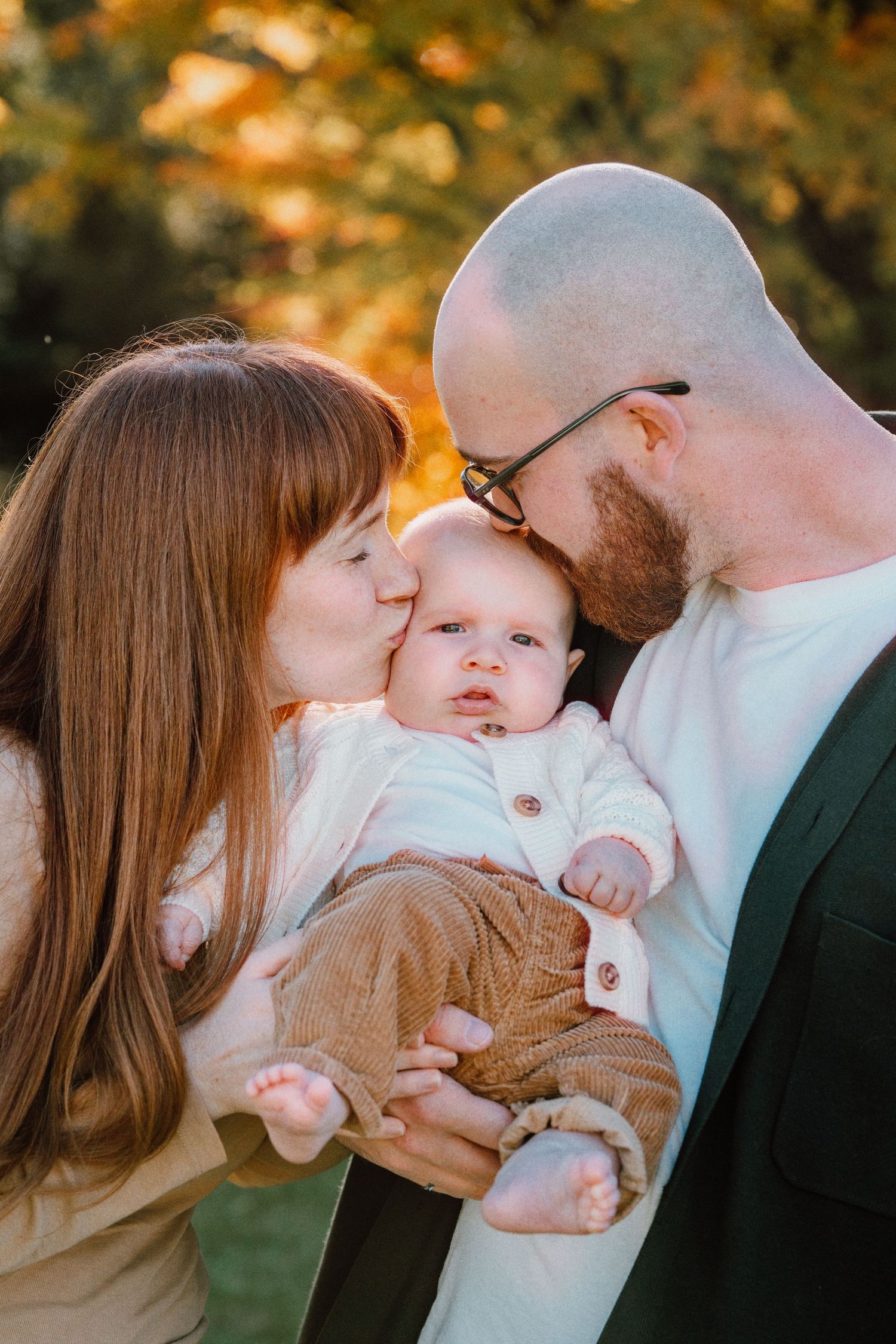 Parents kissing their newborn baby on the cheeks outdoors in autumn park with golden foliage background.