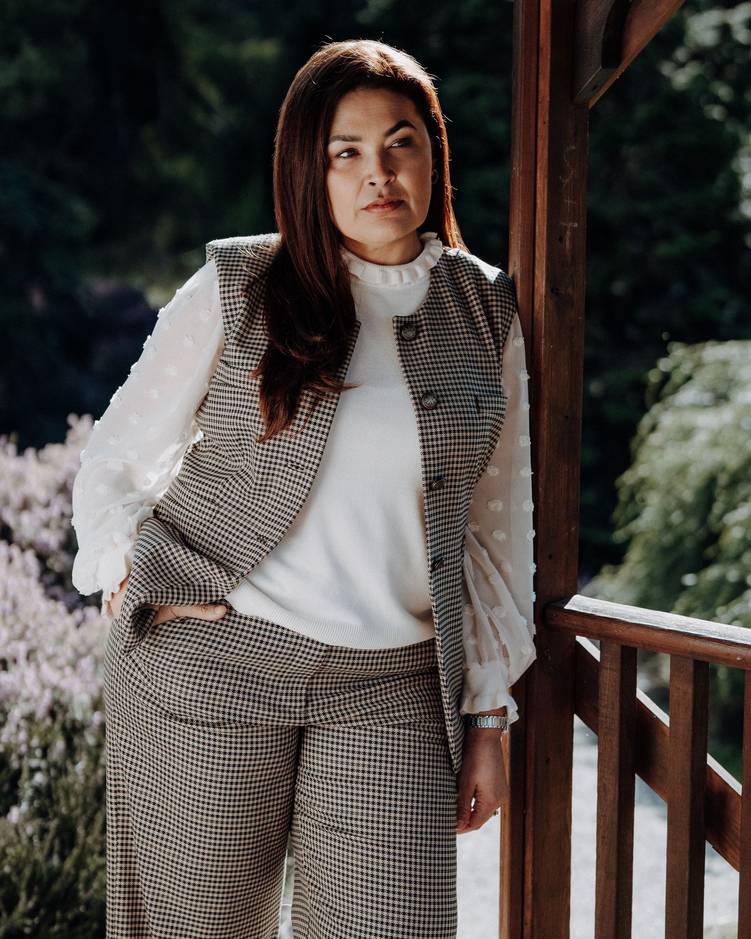 Woman in houndstooth vest suit and white blouse posing outdoors near wooden structure with garden background. Portrait photography