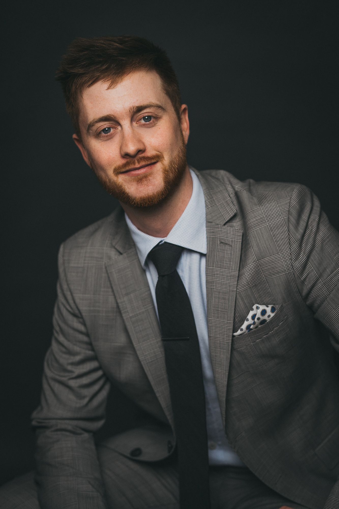 Young man with red beard in gray plaid suit, black tie, and pocket square smiling confidently against dark background.