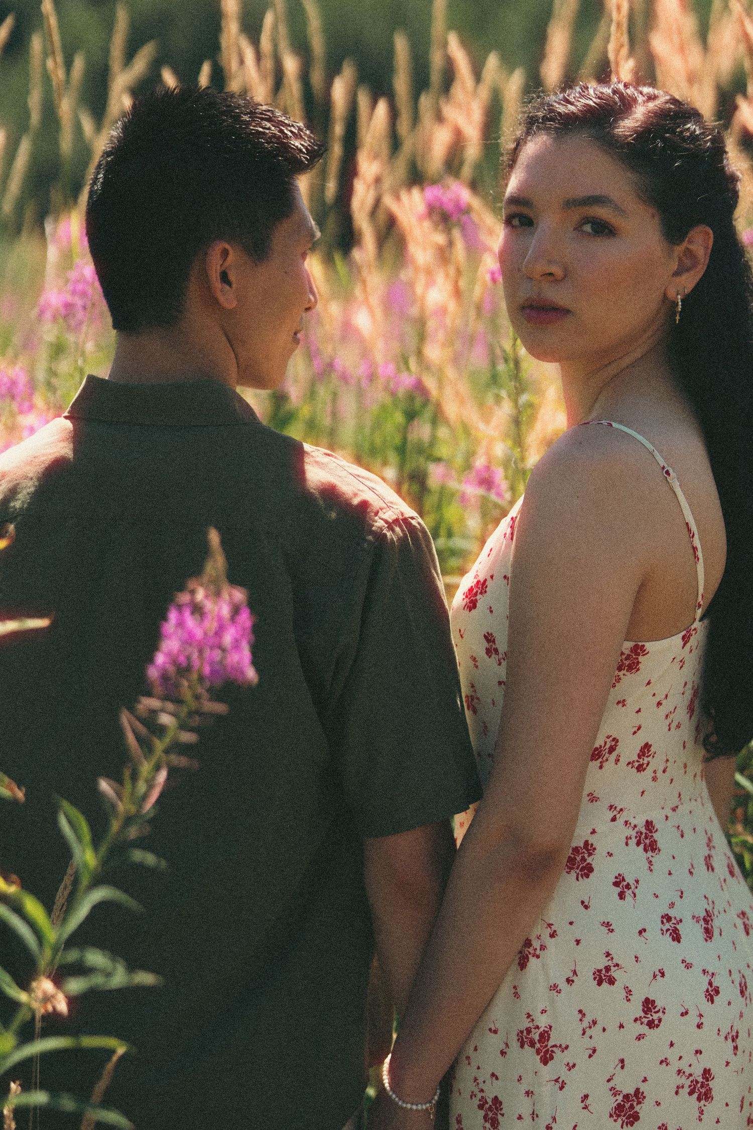 Couple holding hands in a field of pink wildflowers, woman in floral dress looking back at camera.