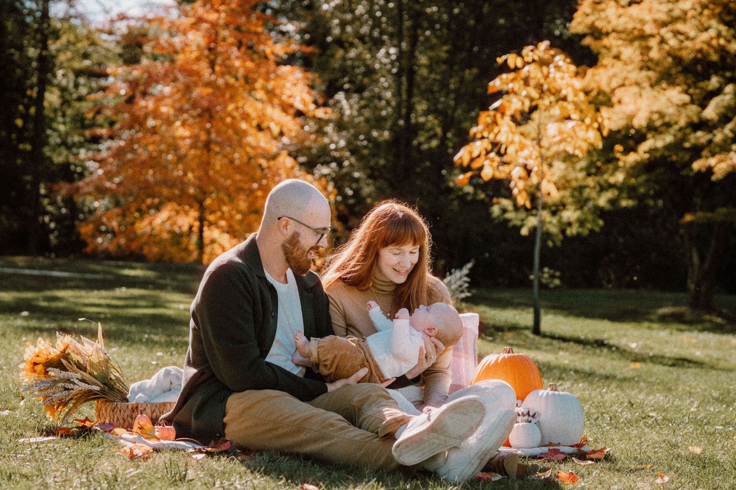 Family sitting on blanket in autumn park with orange pumpkins and colorful fall foliage trees in background.