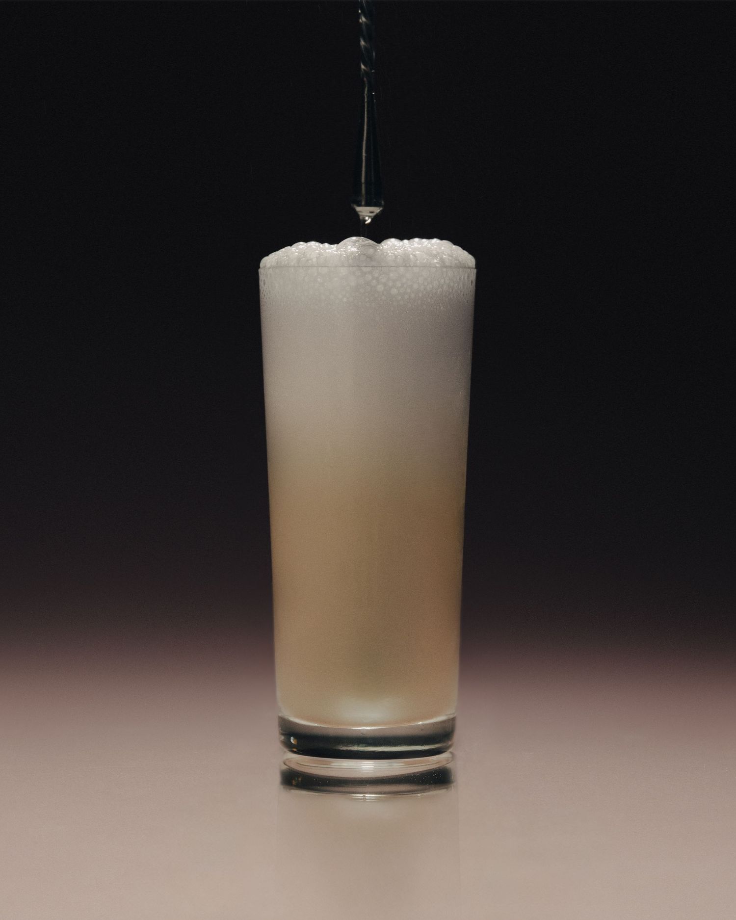 A tall highball glass filled with a frothy, creamy white cocktail being poured against a dark background.