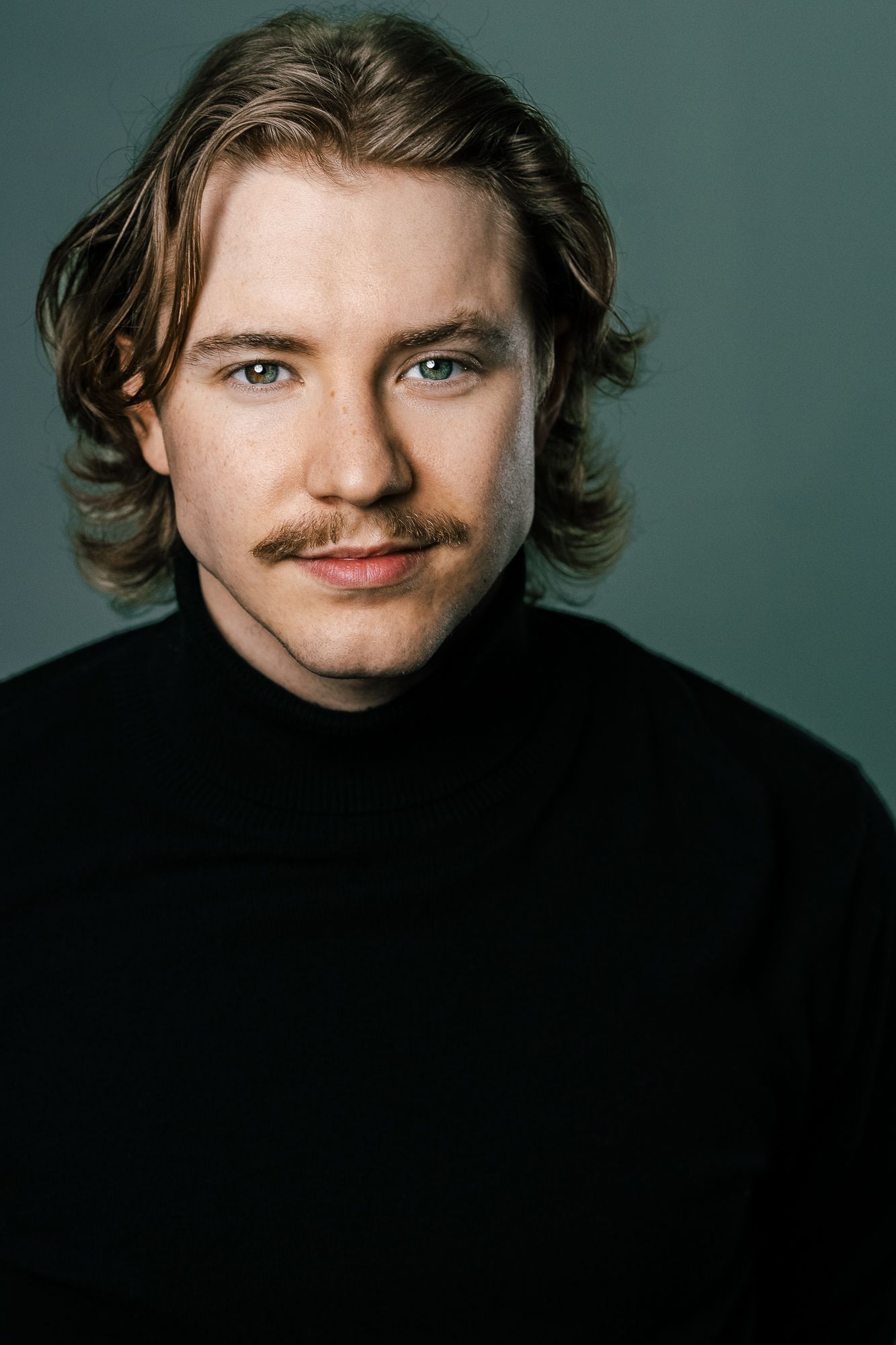 Young man with blonde hair and mustache wearing a black turtleneck sweater, posing for a studio portrait.