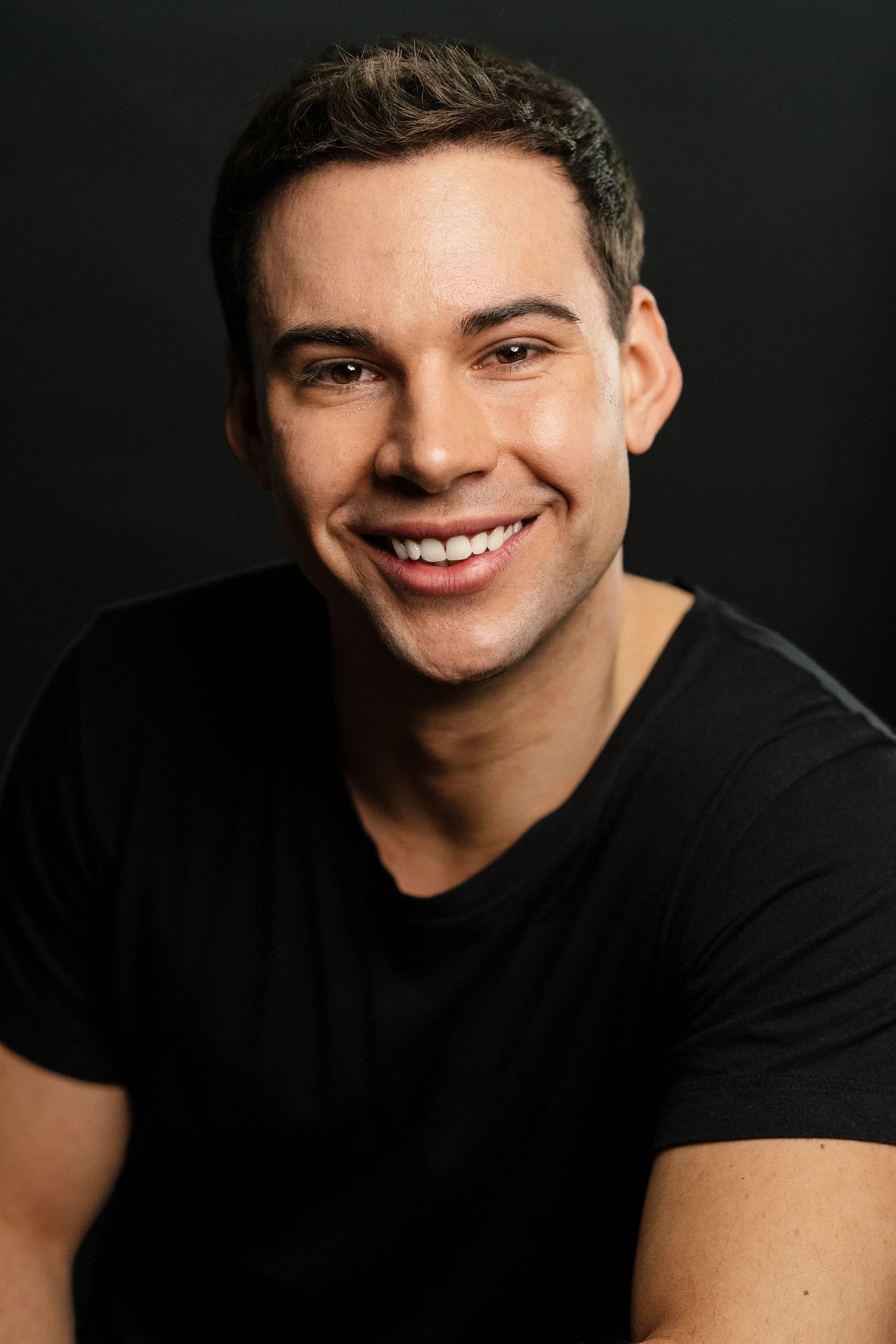 Smiling young man with dark hair wearing a black t-shirt against a dark background in a professional headshot.