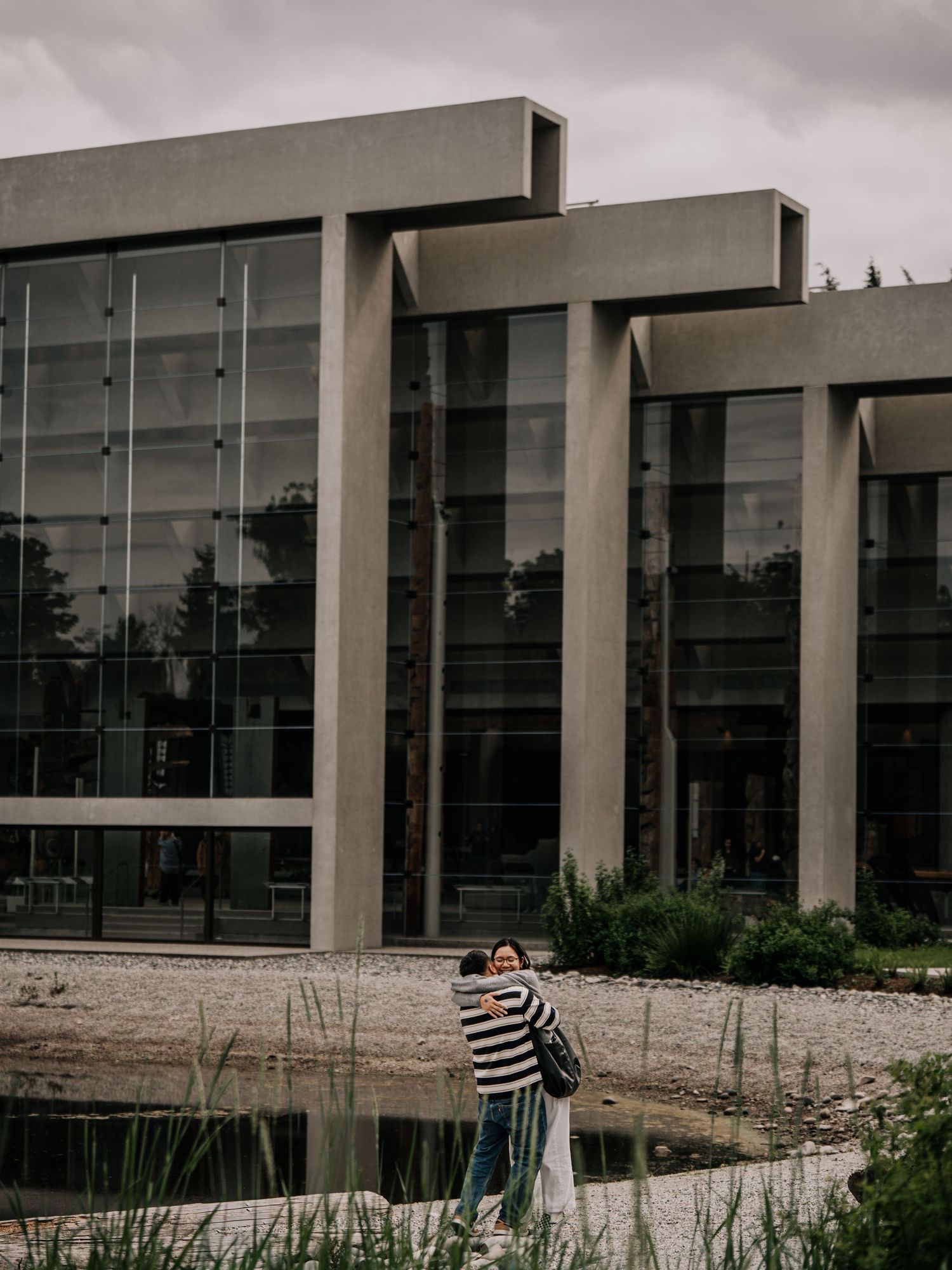 Couple embracing in front of a modern glass and concrete building near a pond.