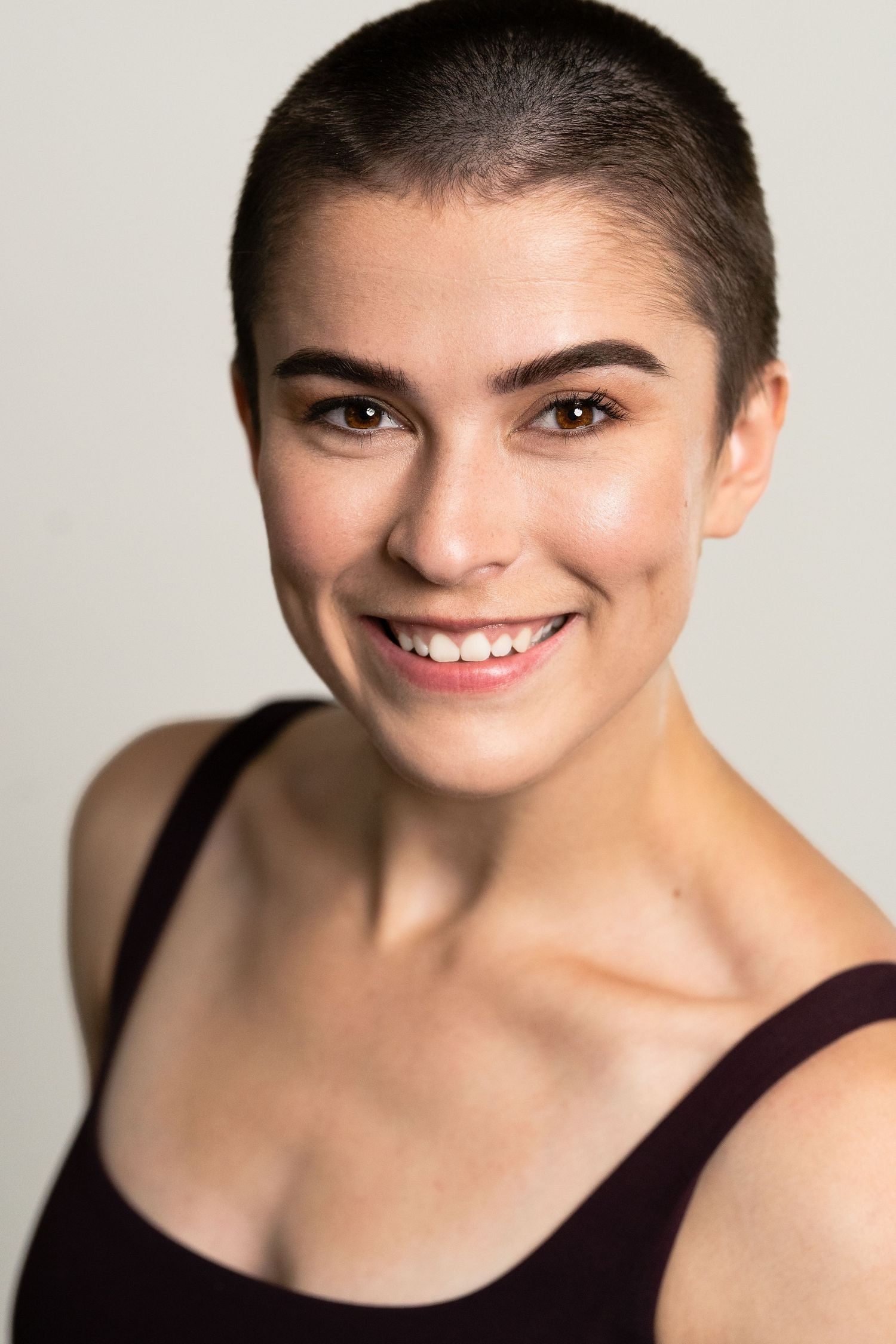 Young woman with short buzz cut hair smiling confidently at camera, wearing dark tank top against light background.