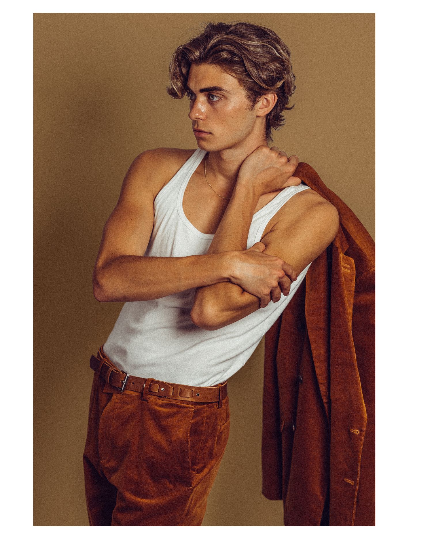 Young male model in white tank top and rust corduroy pants, holding brown jacket over shoulder against tan background. Fashion photo