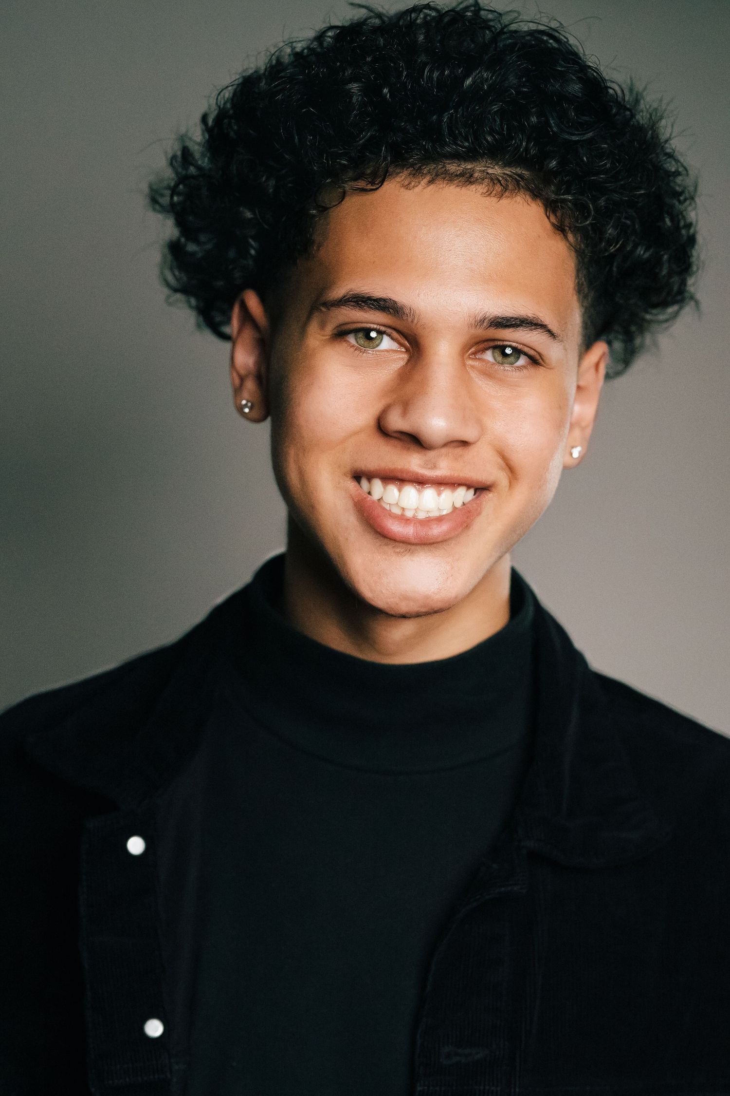 Young man with curly black hair, ear studs, and a bright smile wearing a black turtleneck and jacket.