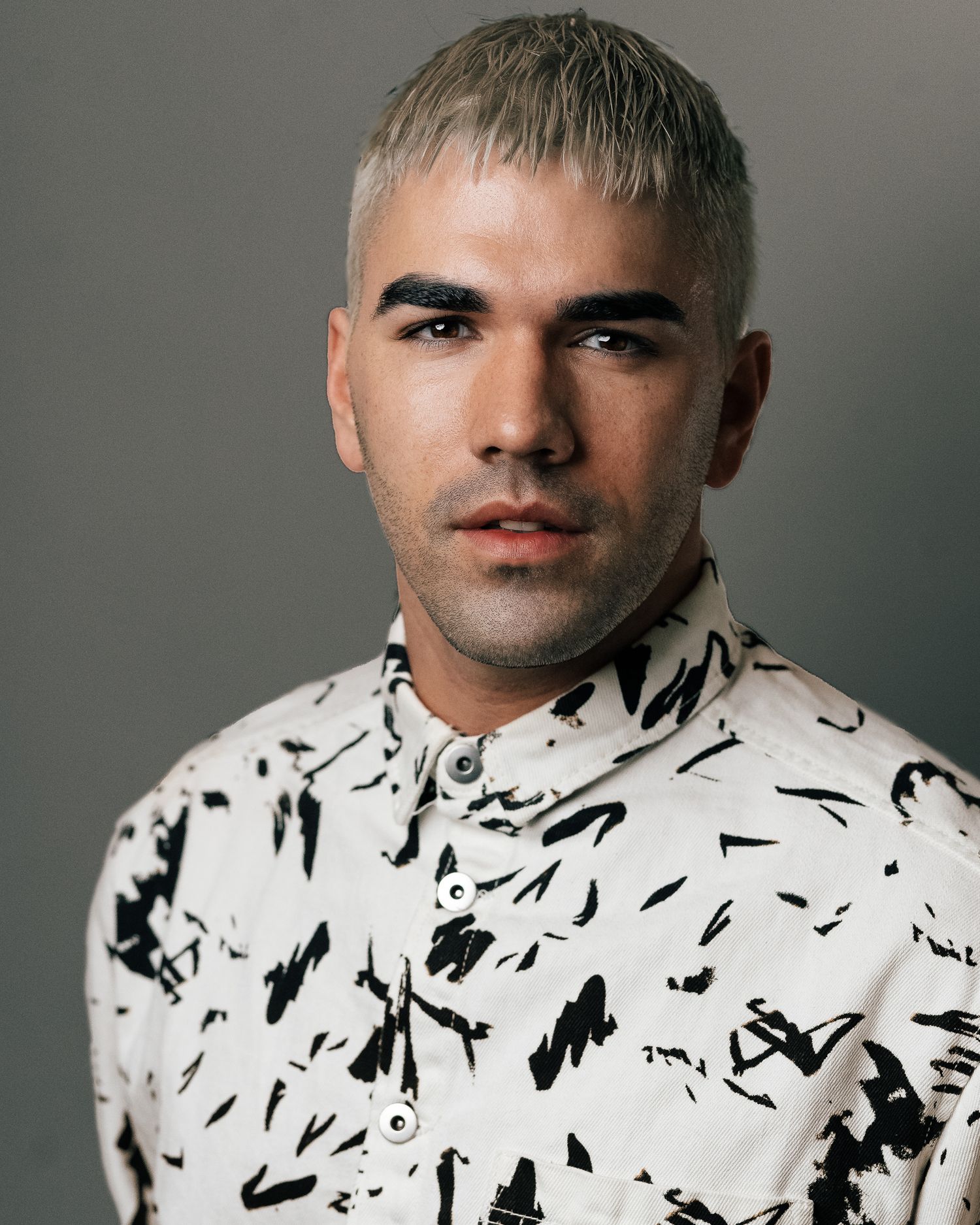 Man with bleached blonde hair wearing a white and black abstract print shirt poses for a portrait photo.