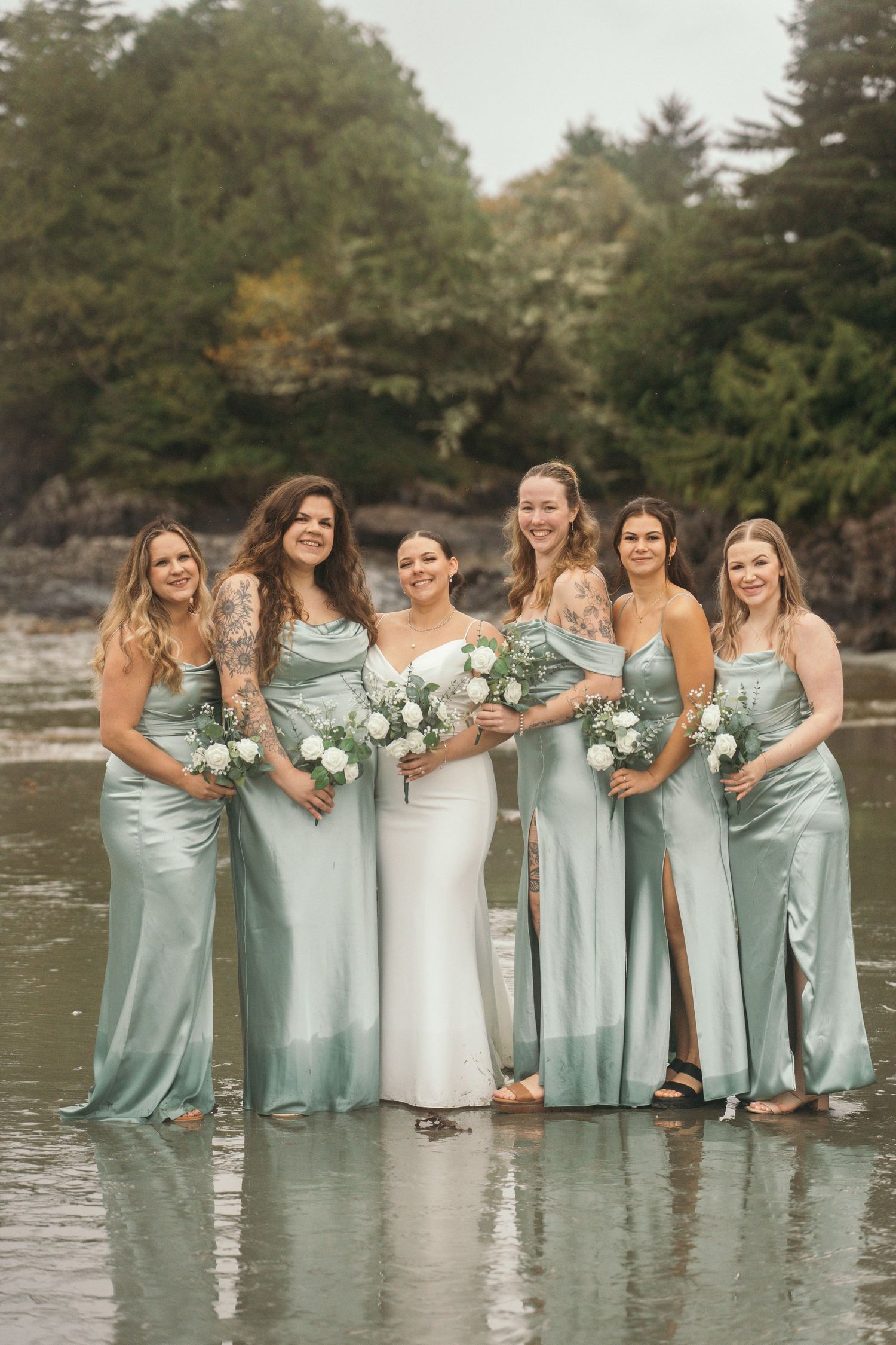Bride in white gown with five bridesmaids in sage green satin dresses holding white bouquets on a beach.