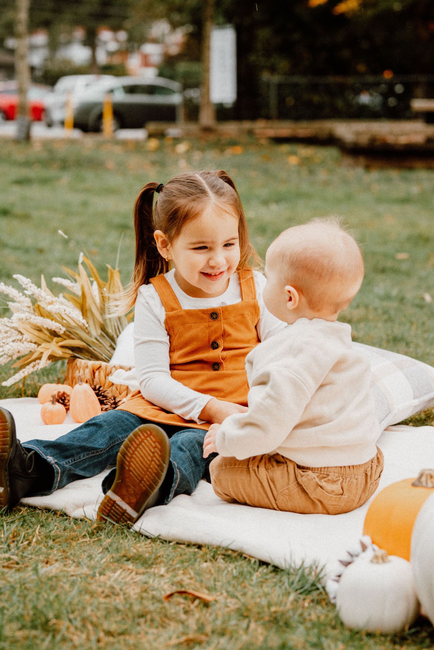 Two young children sit on a blanket outdoors in fall, surrounded by pumpkins and autumn decor.