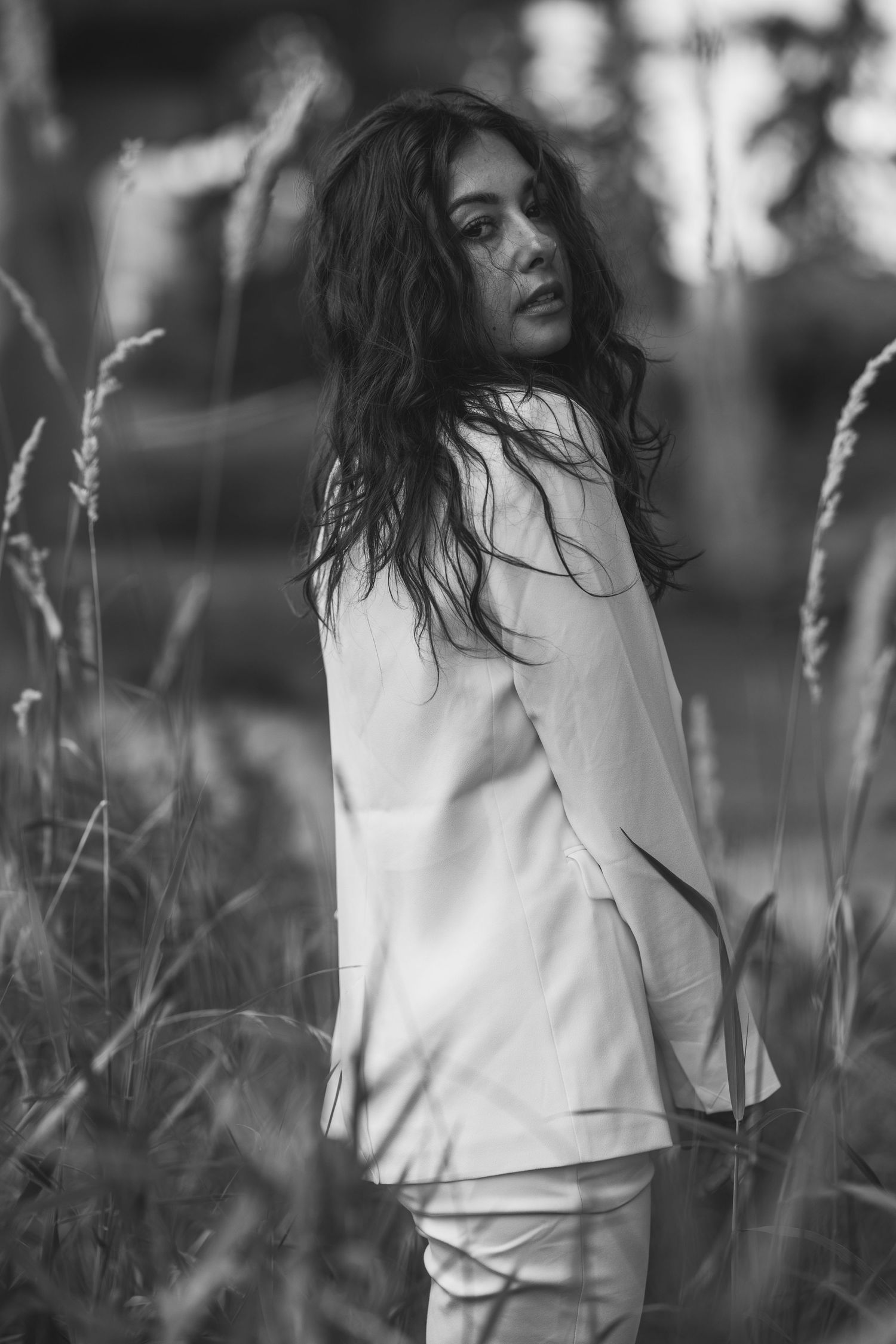 Black and white portrait of a woman with long hair in a white suit standing among tall grass outdoors. vancouver Lifestyle photography