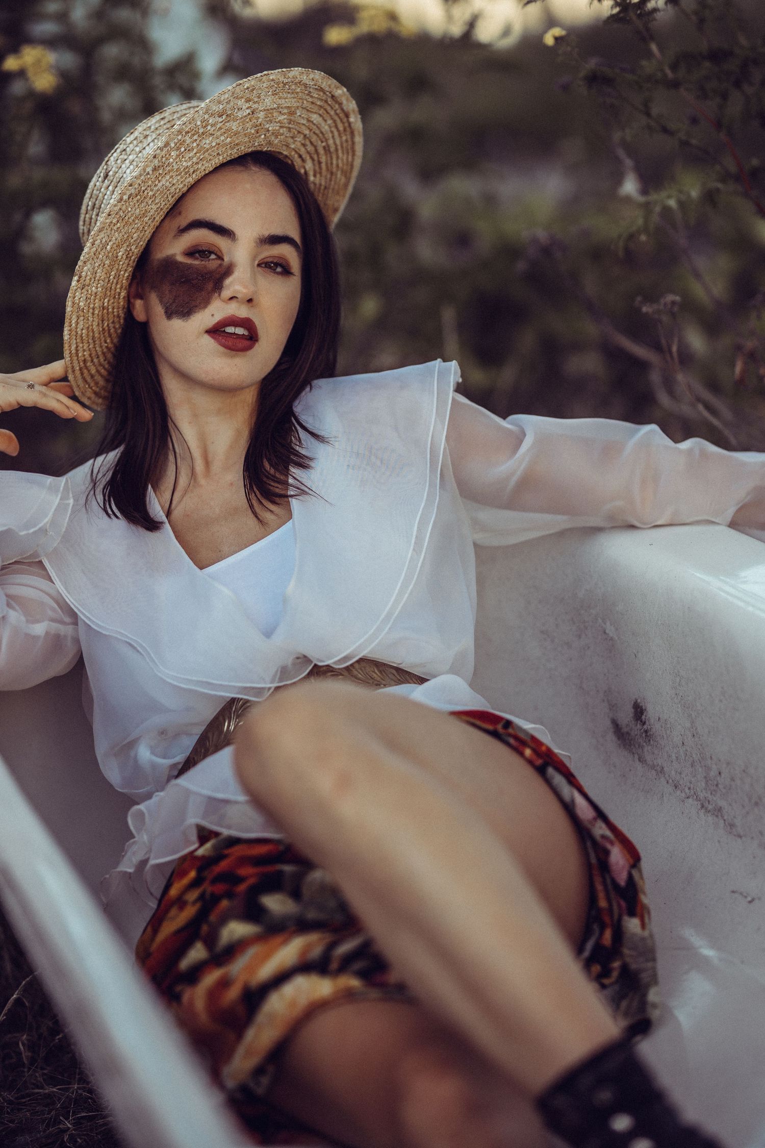 Young woman in straw hat and white sheer blouse relaxing in outdoor bathtub surrounded by nature.