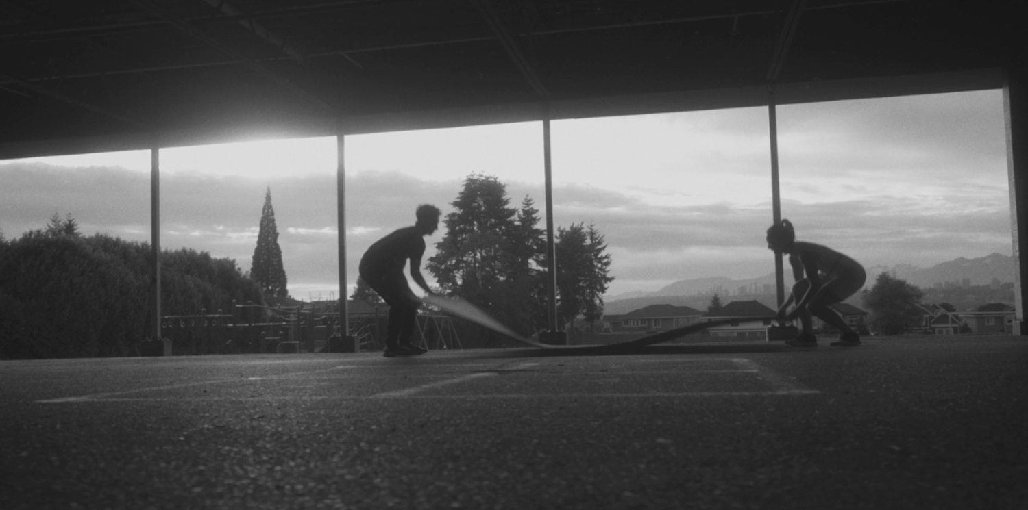 Two athletes battle ropes in dramatic black and white silhouette under an open structure with mountain skyline backdrop.