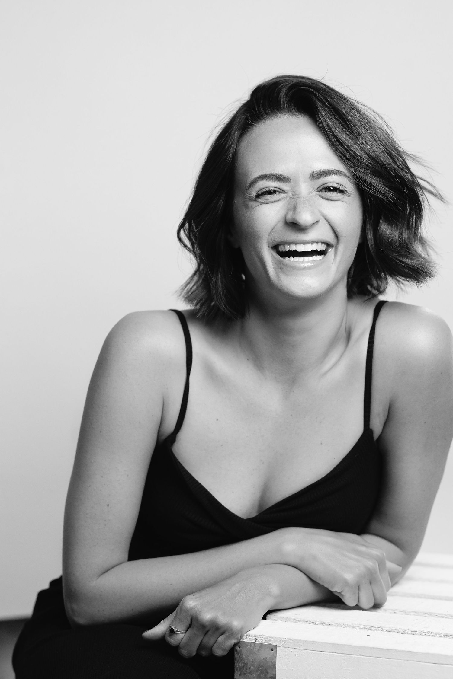 Black and white portrait of a laughing woman in a black dress, leaning on a white surface.