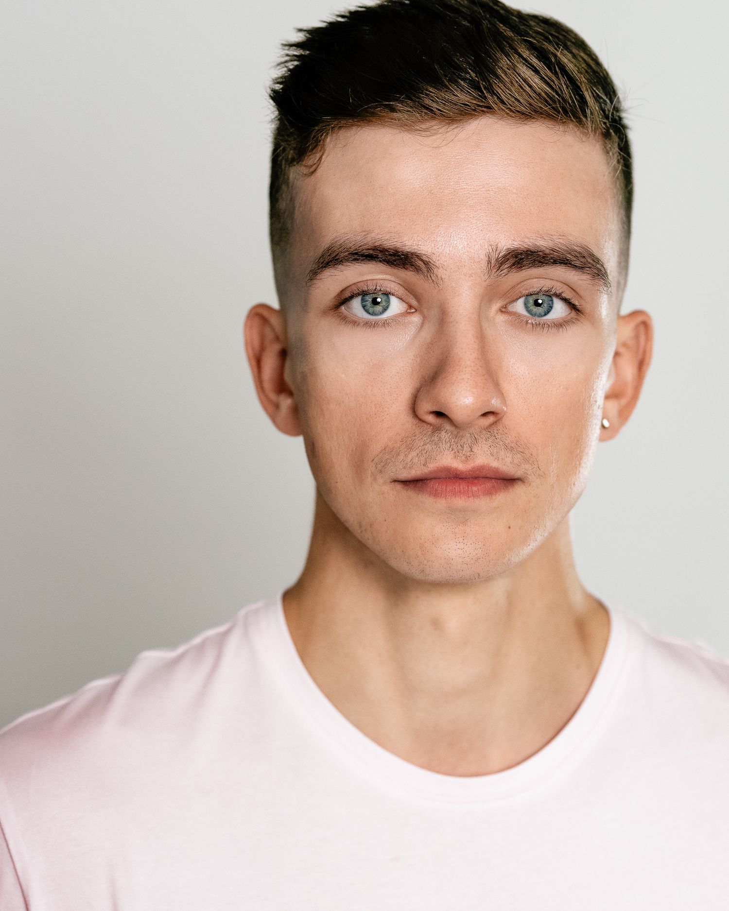Young man with blue eyes, short brown hair, and a small earring wearing a light pink t-shirt against a gray background.
