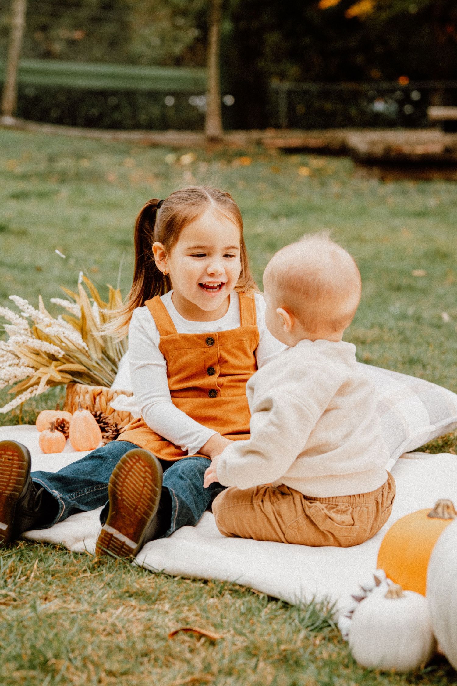 Two young children sit on a blanket outdoors during fall, surrounded by pumpkins and dried flowers.